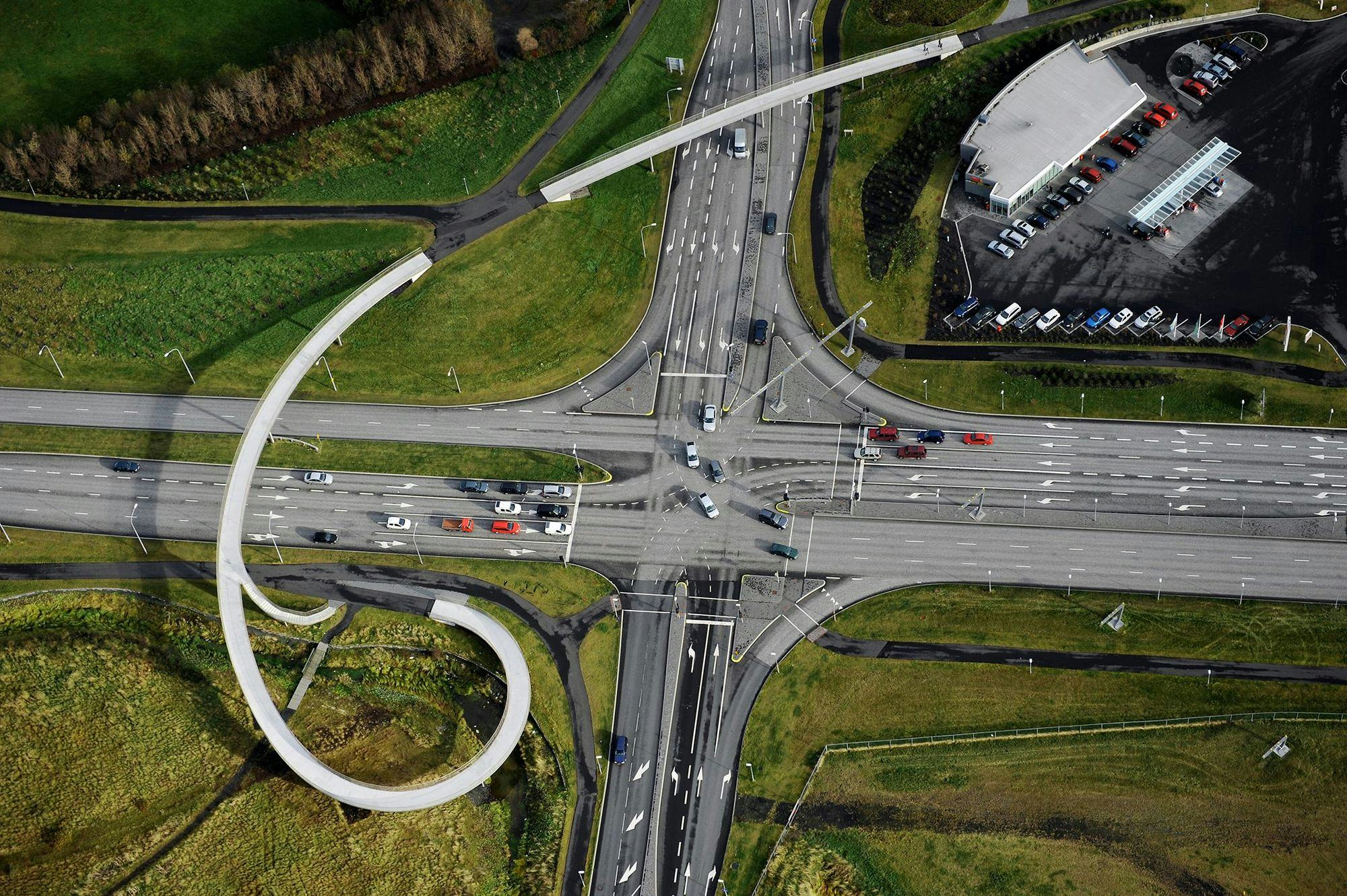 A central intersection is visible from the air and down, we look down on traffic streets that cross, a pedestrian bridge crosses the street on one side, cars on the streets and houses on the other side of the street.