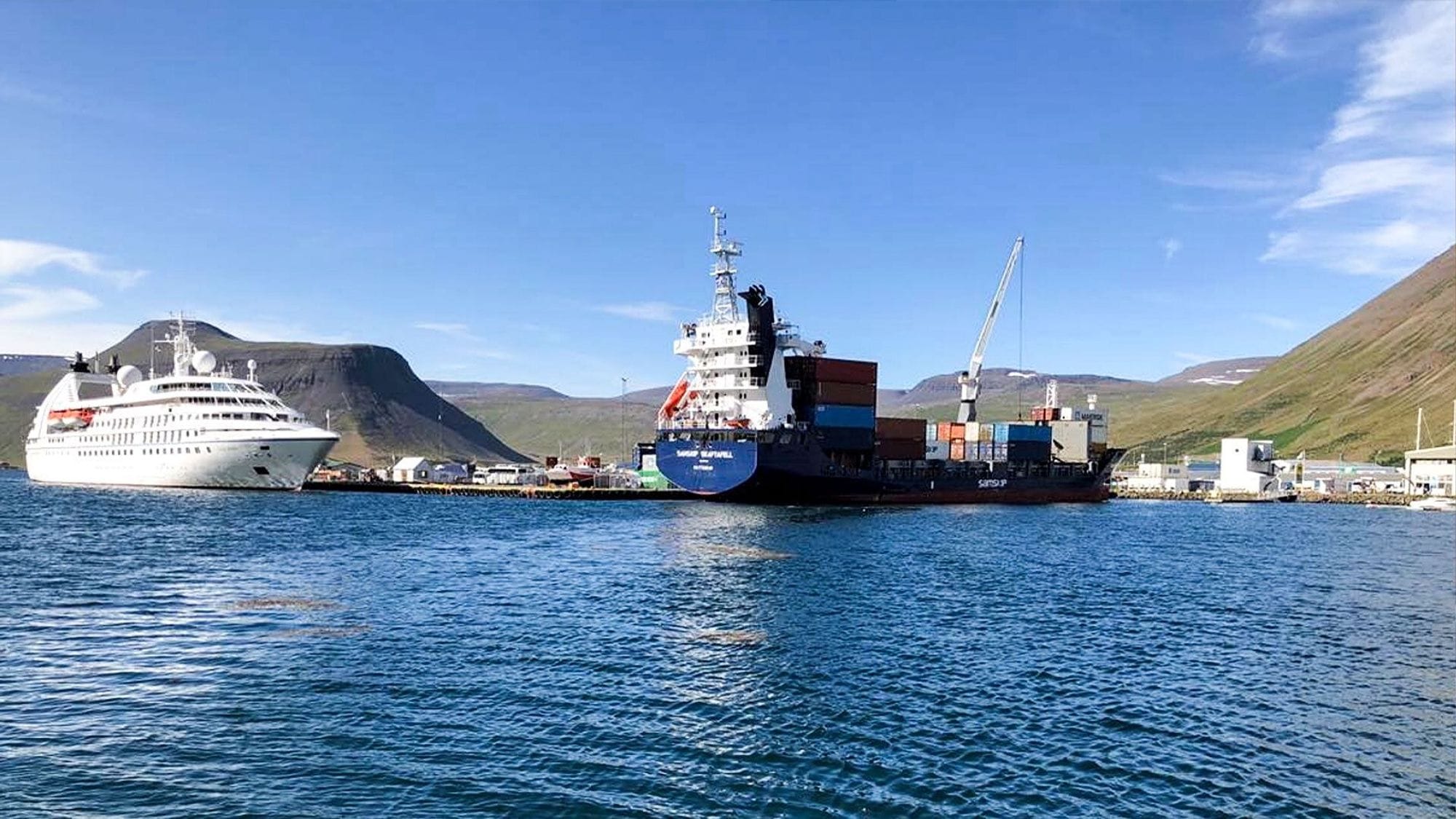 Horft af sjó í átt til hafnar, landslag í bakgrunni, stórir togarar, skemmtiferðaskip og gámasvæði á höfninni