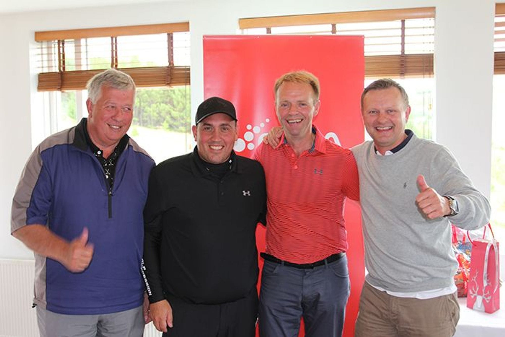 Four smiling men giving thumbs up in front of a red banner