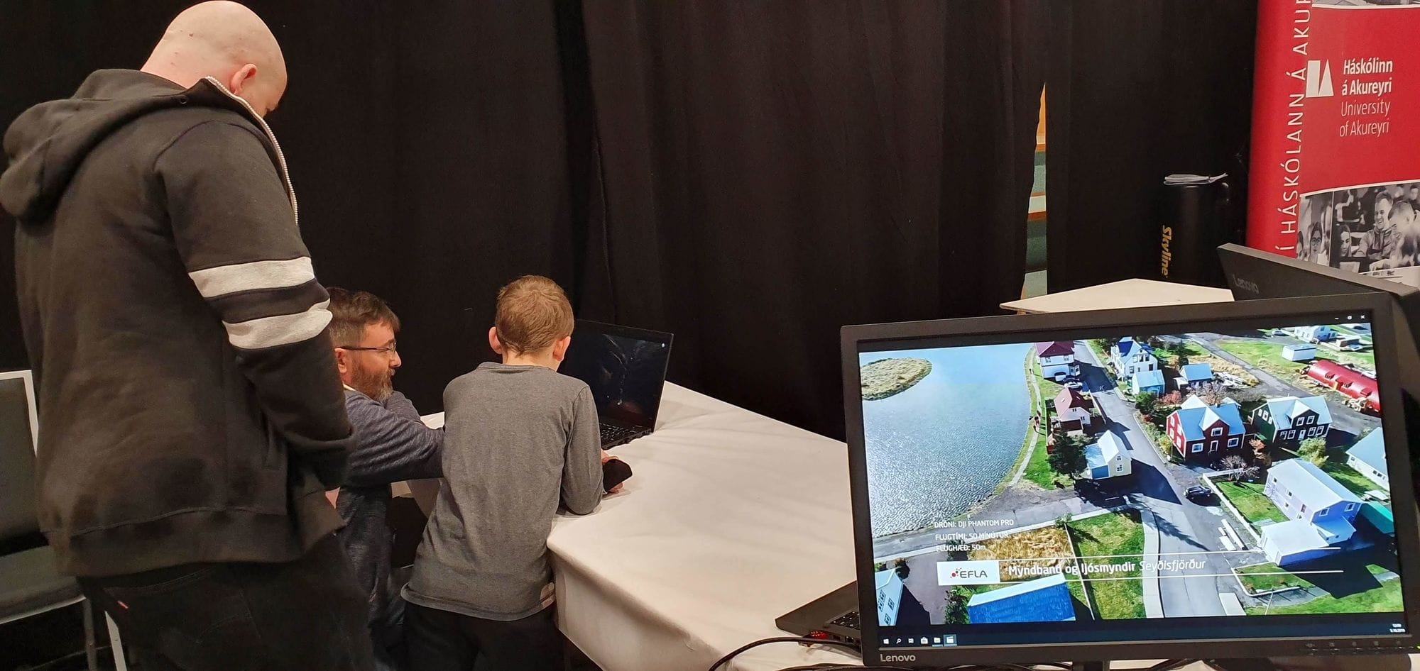 Two men and a child looking at a laptop screen and a laptop screen displaying aerial drone footage at the right bottom corner