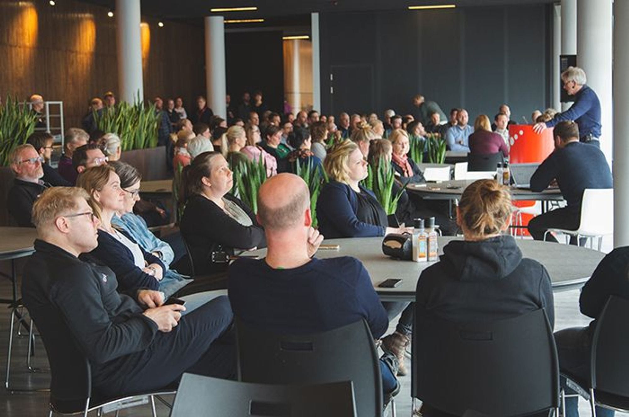 People seated at round table focused on speaker standing in the center of the room