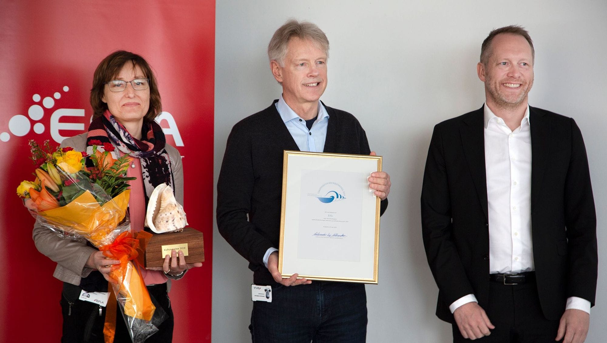 Three individuals posing with smile on their faces, holding framed certificate and awards