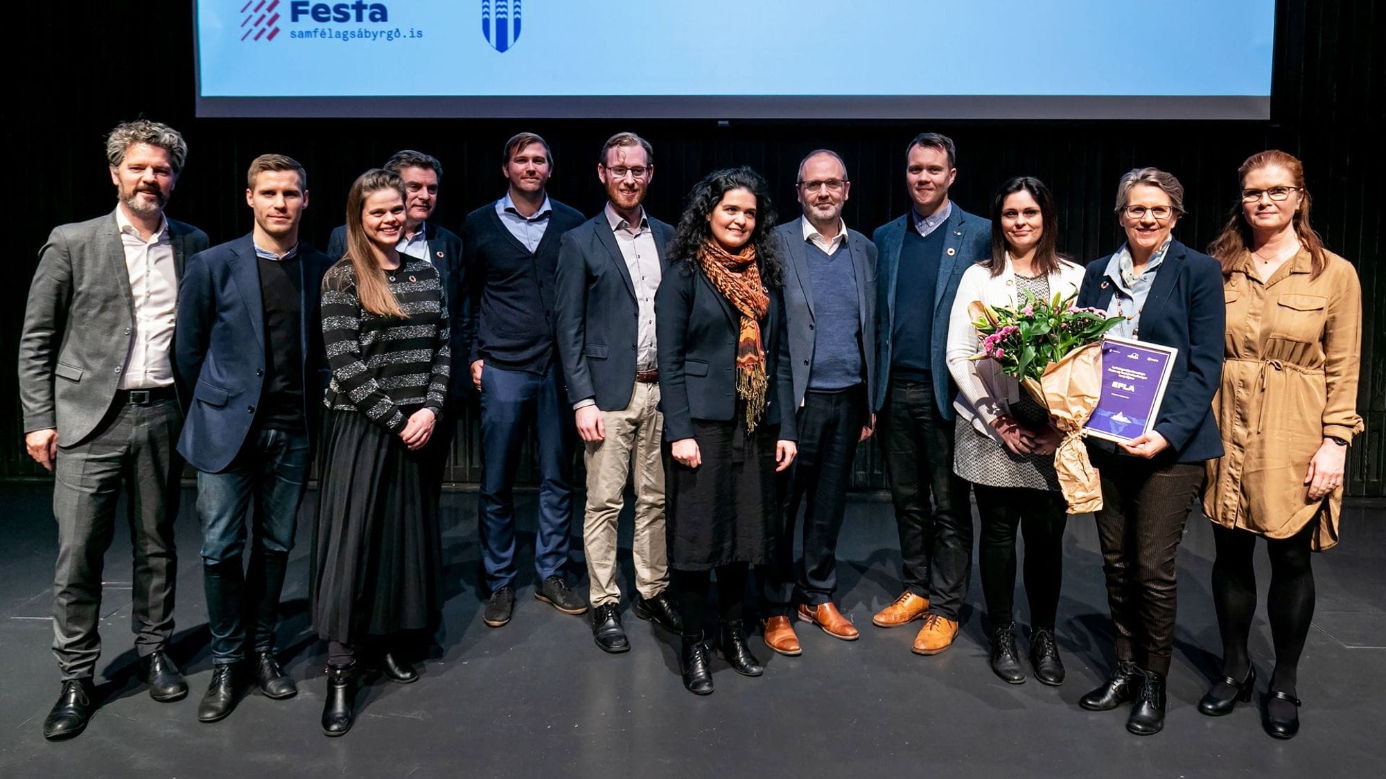 A large group of people standing together posing with a smile on their faces and two women from the group are holding certificate and bouquet 