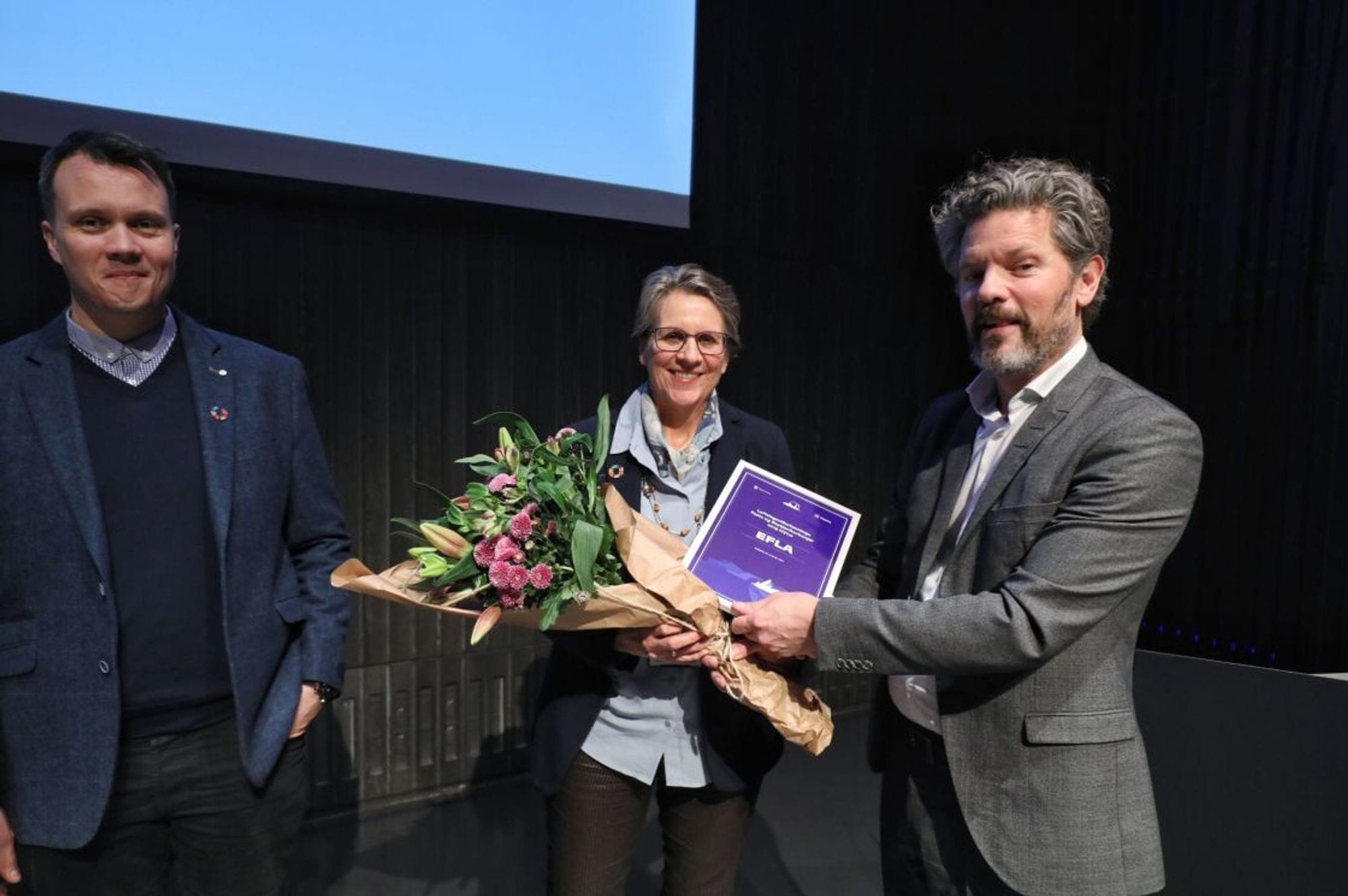 A man in grey suit presenting bouquet of flower and a certificate to a woman, next to a man in blue suit