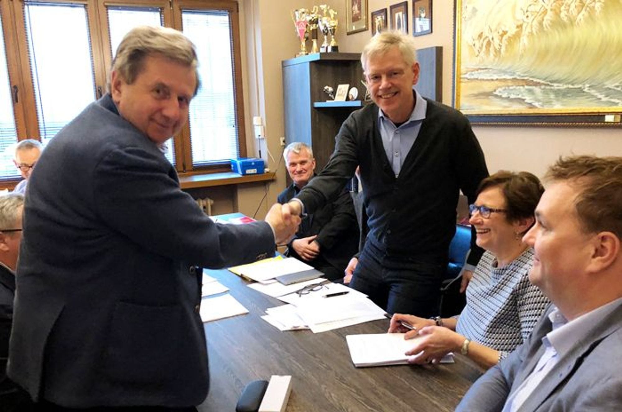 Two men shaking hands and few other people sitting and looking at them 