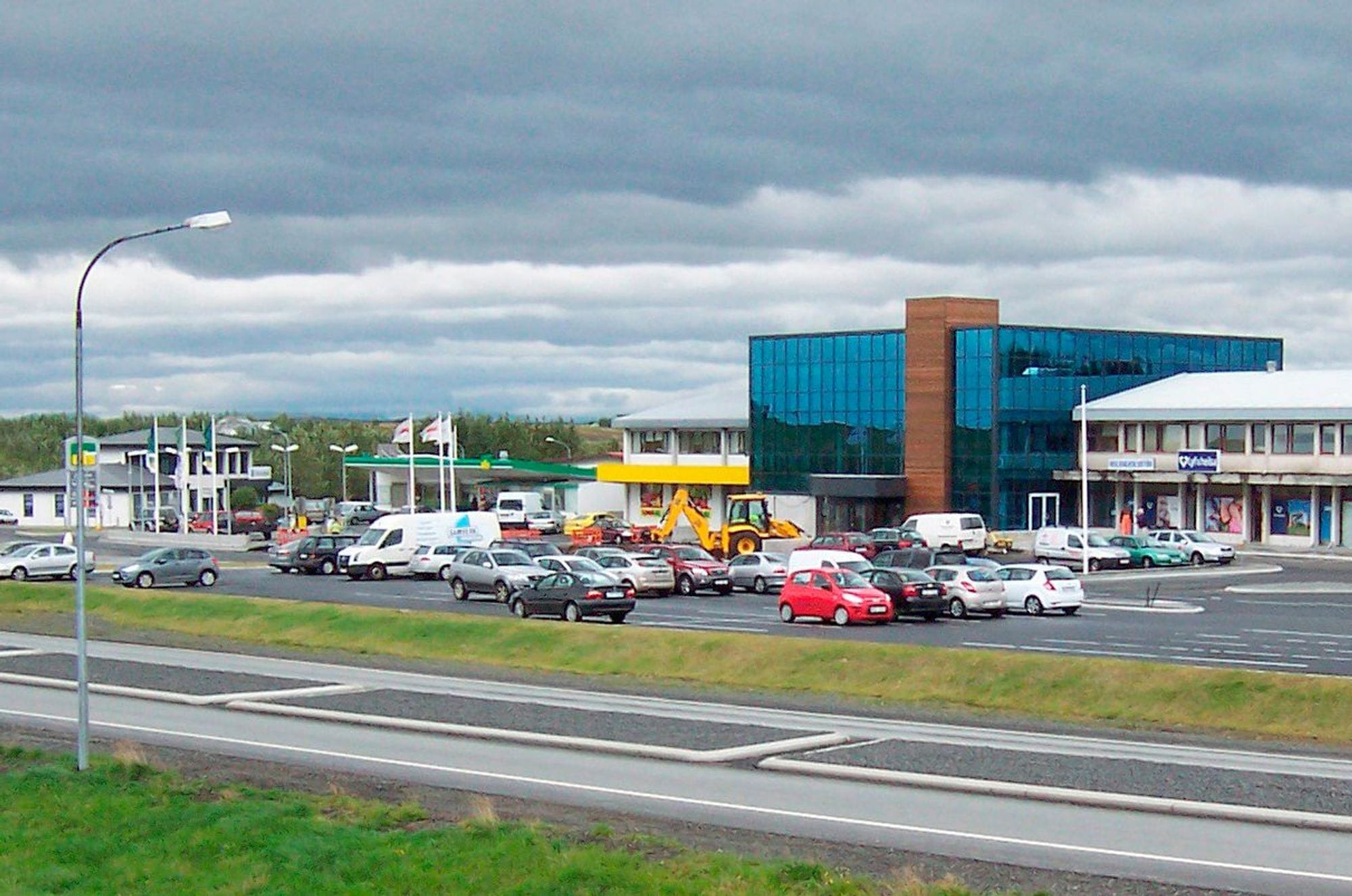 Parking lots with cars in front of dark blue glass building, two story white buildings and a gas station