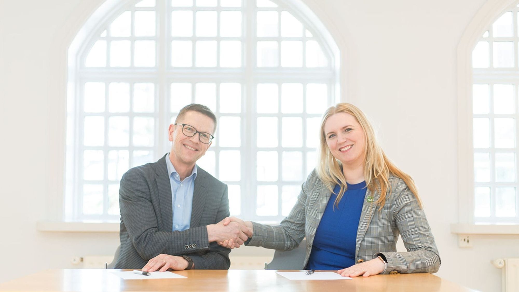 A man and a woman in formal attire smiling and shaking hands across a table featuring a grid patterned window in the background