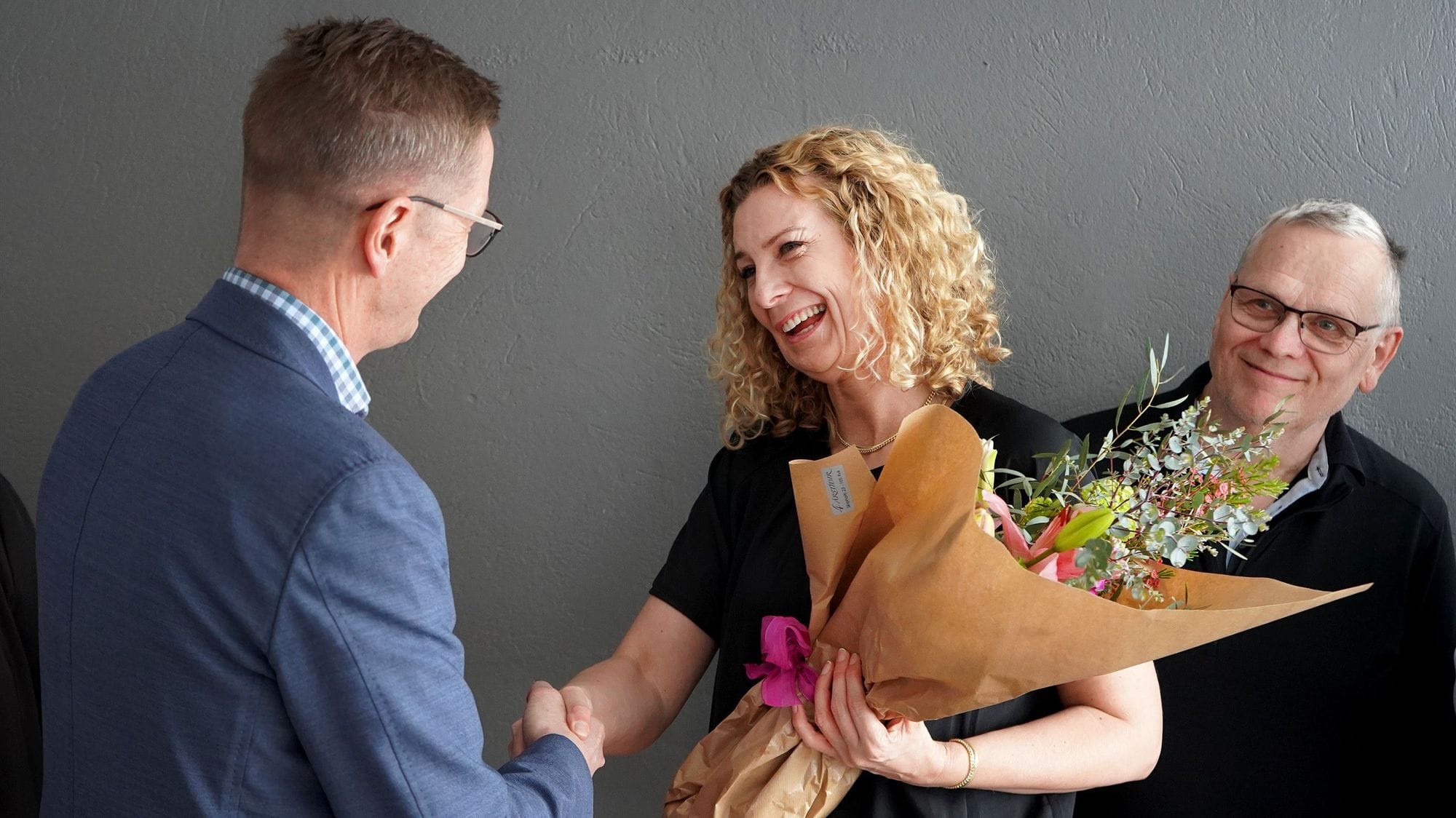 A man and a woman shaking hands with the woman holding a bouquet of flowers and another person smiling in the background