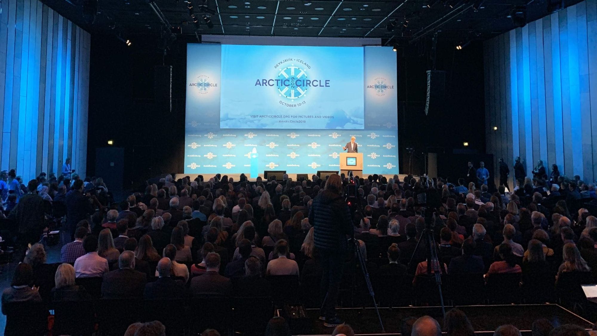 A man delivering speech to an audience with a big screen in the background 