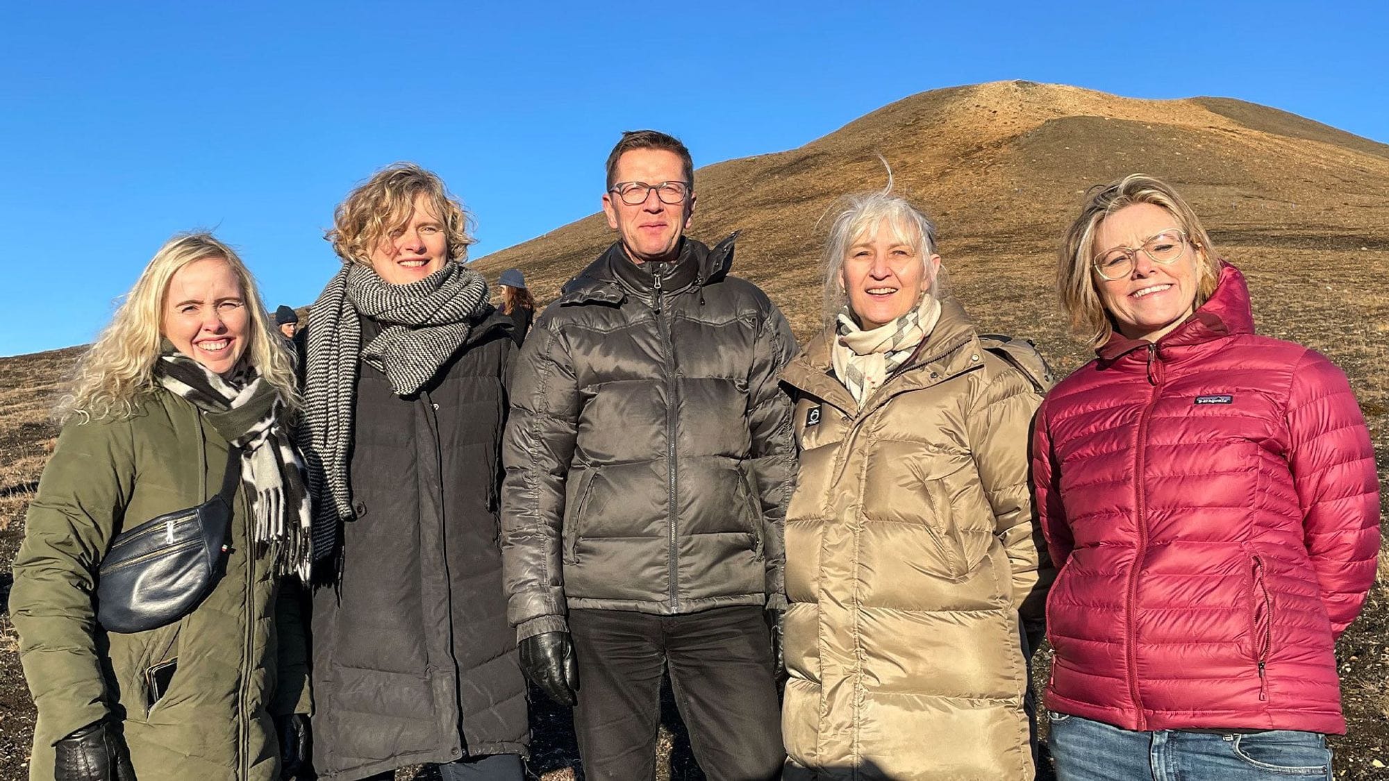 Five individuals dressed in warm clothing, standing outdoors with a barren hill in the background