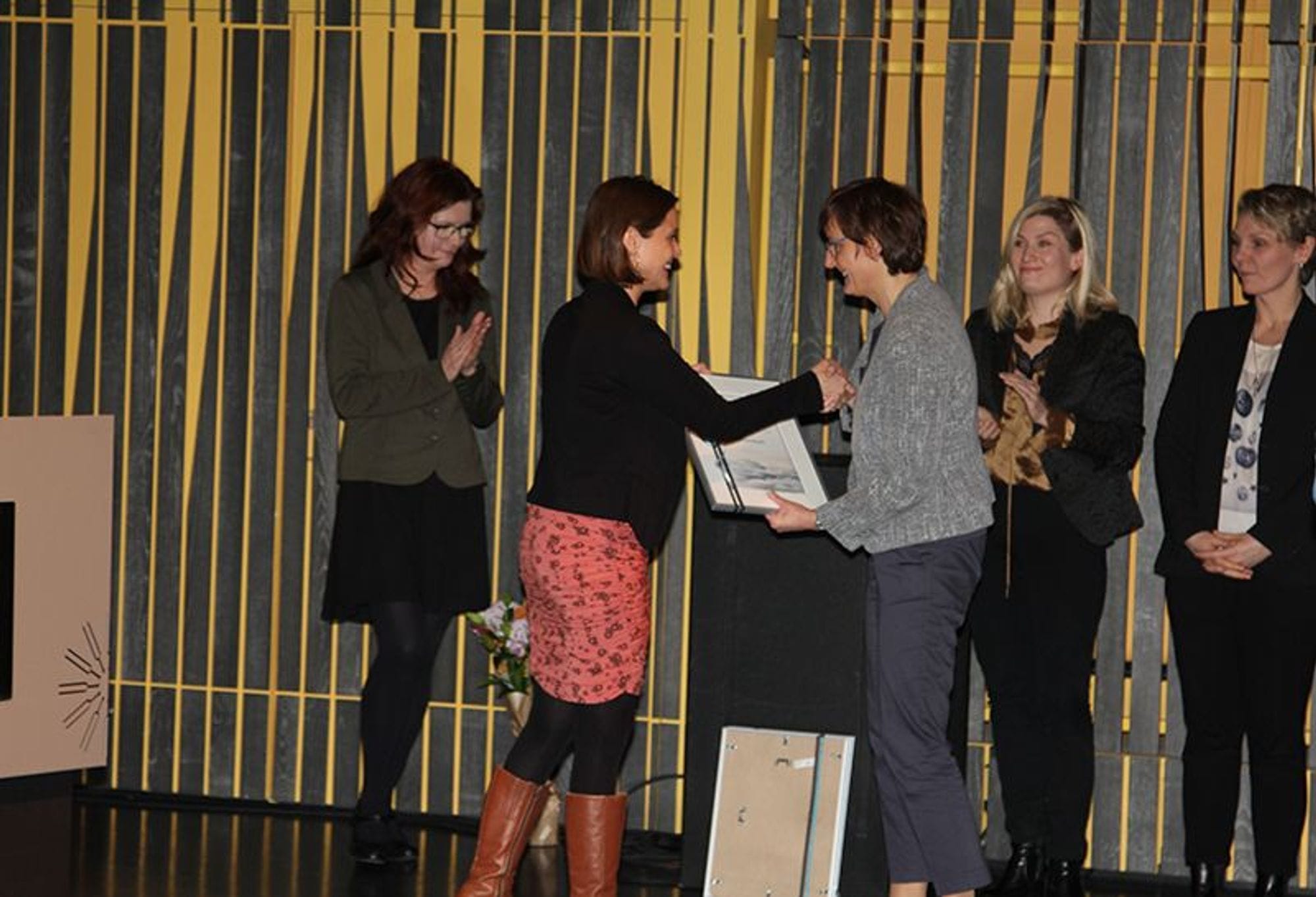 Two women shaking hands and three  women are applauding at the back