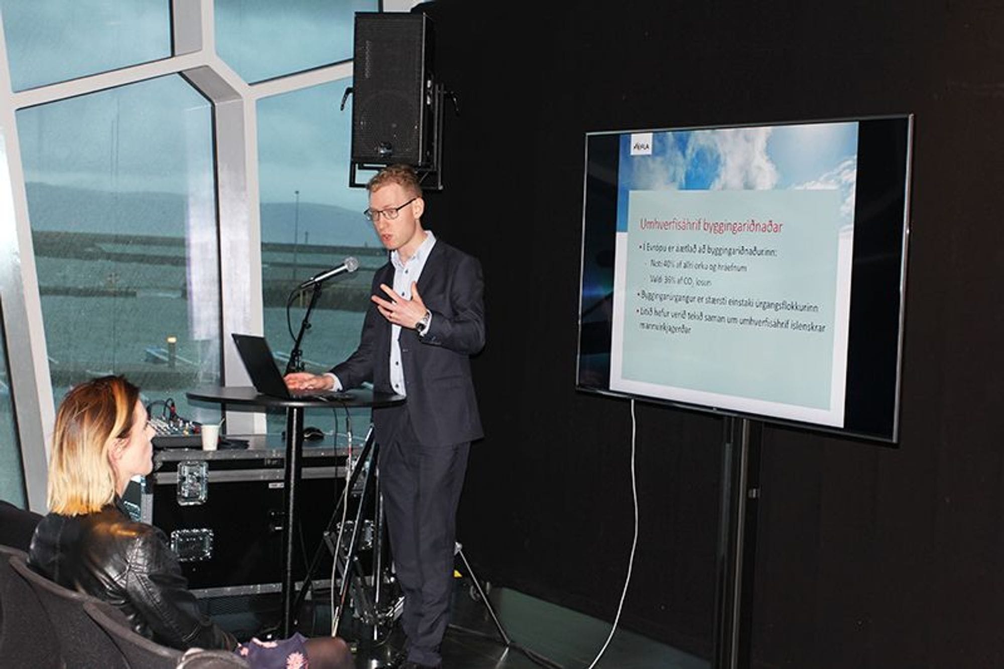 A man giving speech next to a screen with slides and a woman watching them