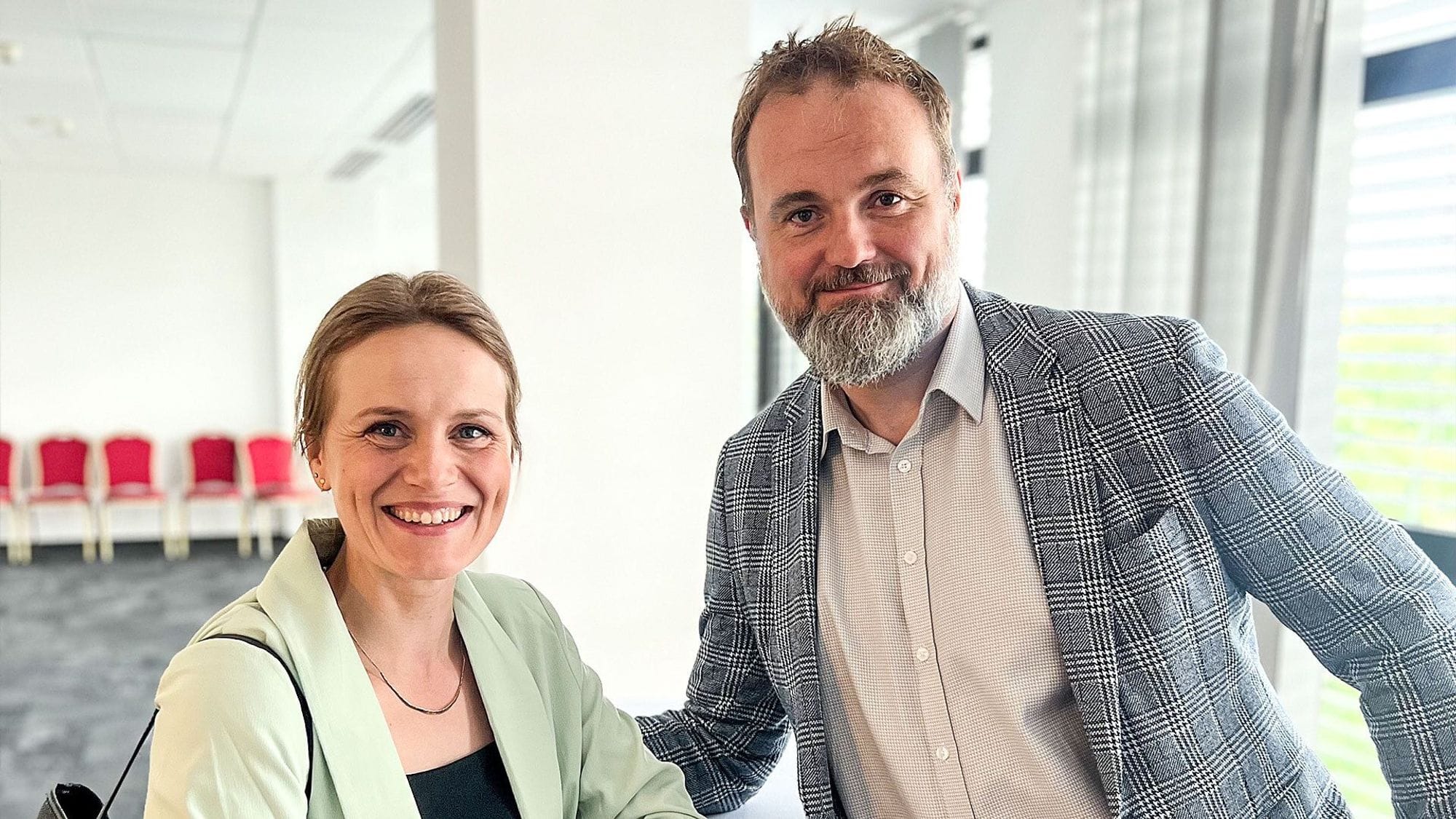 A woman in green blazer and a man in plaid blazer, both smiling towards the camrea