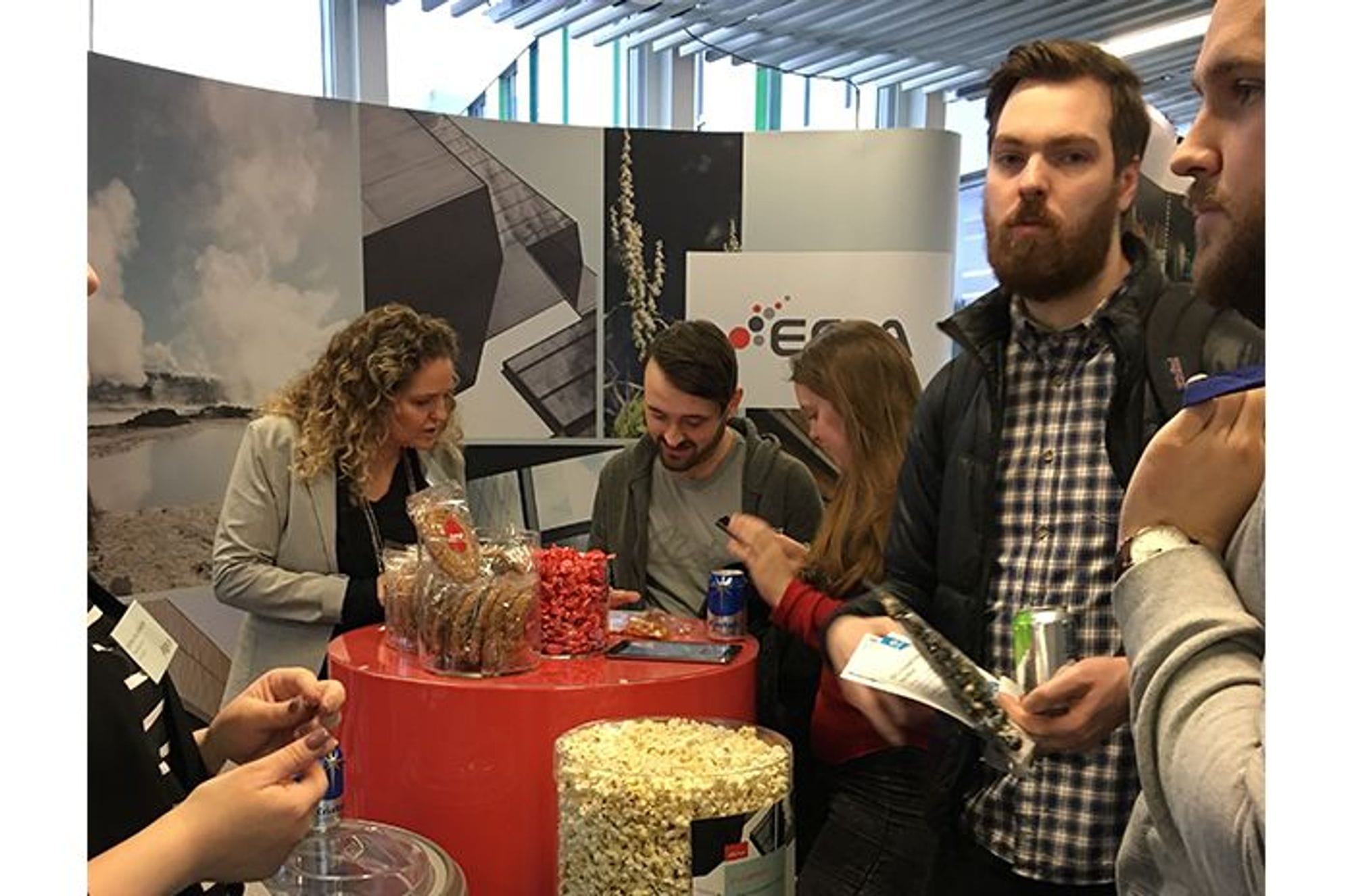 Few people standing around a red round table with snacks and drinks on it