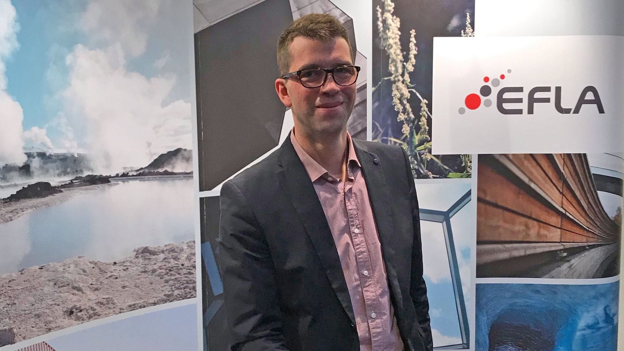 A man in a grey suit, wearing glasses posing with promotional banner in the background