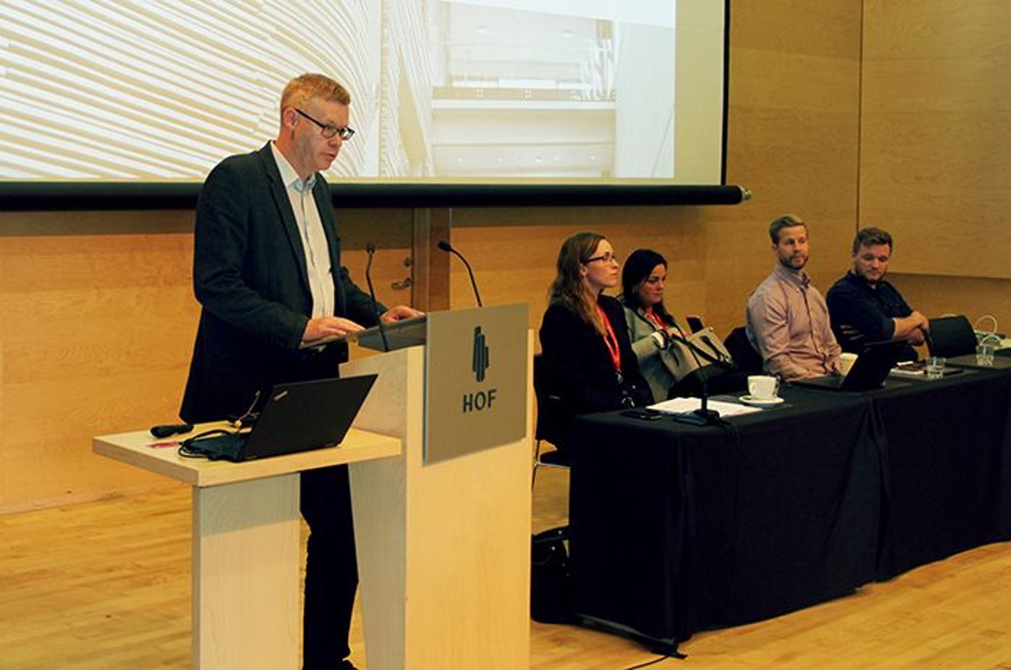 A man  standing  at a podium giving speech and four people seated at a table behind him
