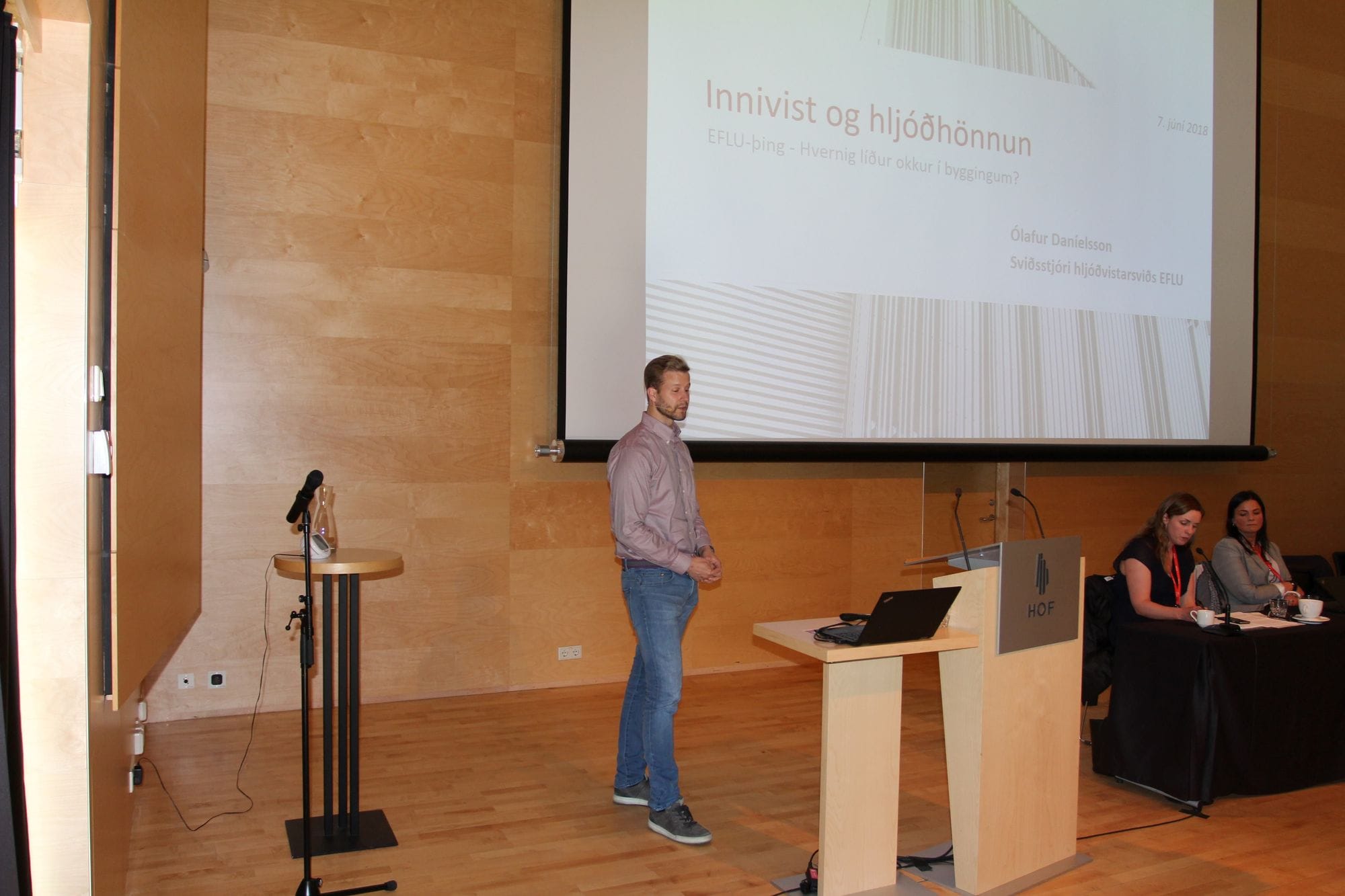 A man giving speech, watching his laptop and two people seated on a table behind the podium