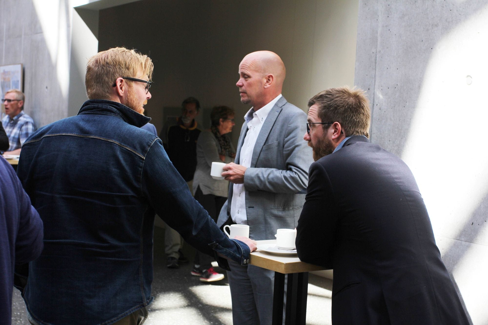 Two men having a conversation and a third man standing between them, holding a cup