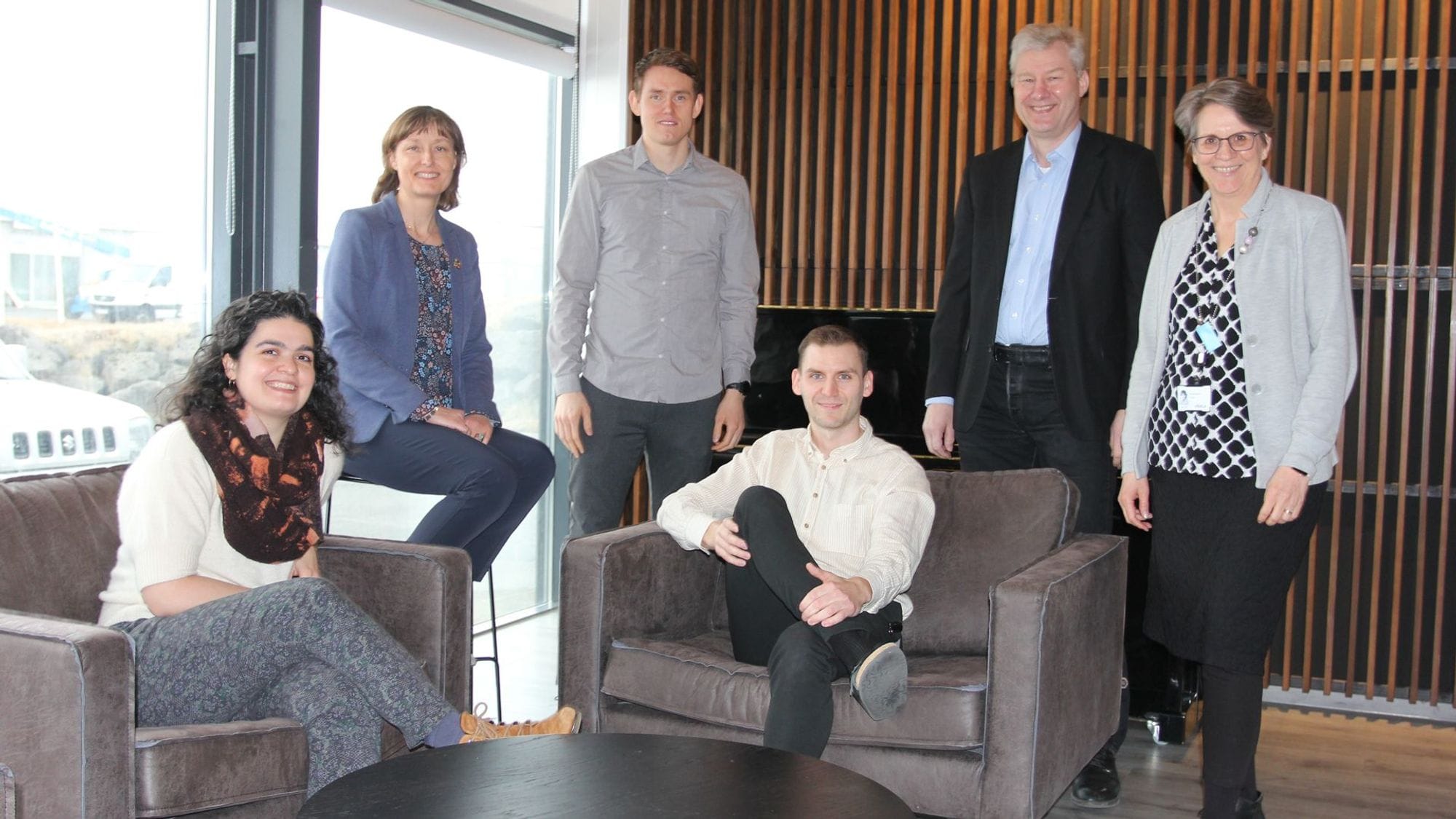 A group of six people, four standing and two seated, posing for a picture in an indoor setting with large windows and modern furnishing
