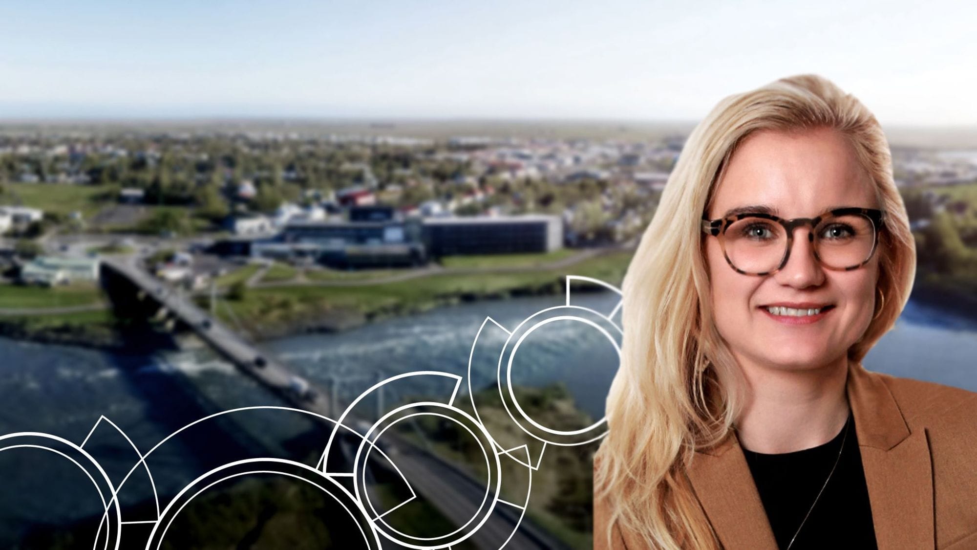 A smiling woman with glasses in the foreground and a blurred background of cityscape by a river