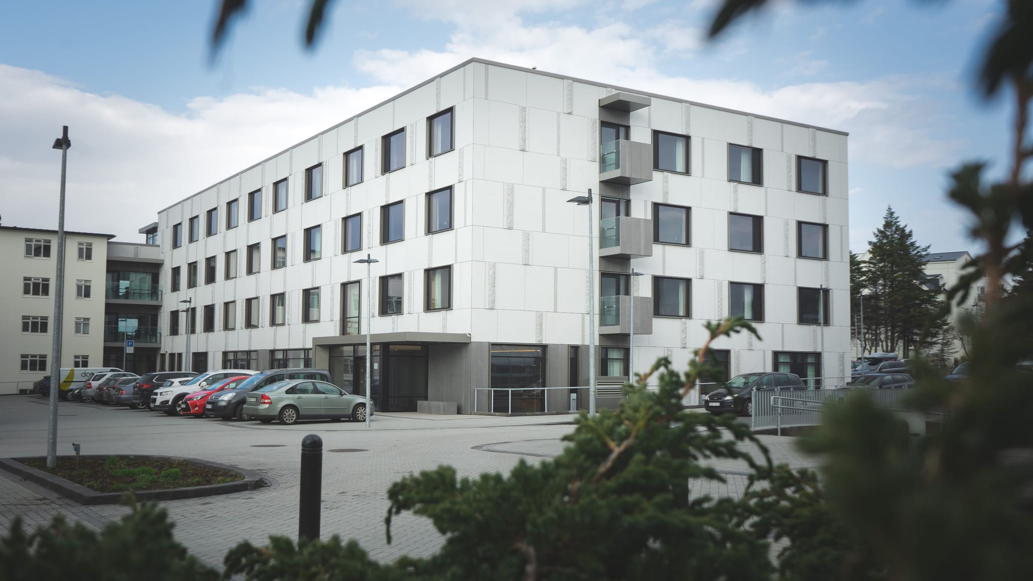 A modern multi-story white building with balcony and parking lot with many cars parked