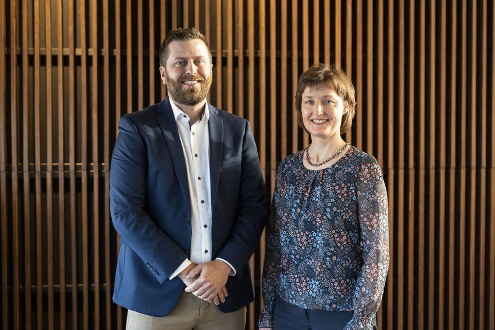 A man and a woman standing with a smile in front of a vertical wooden wall