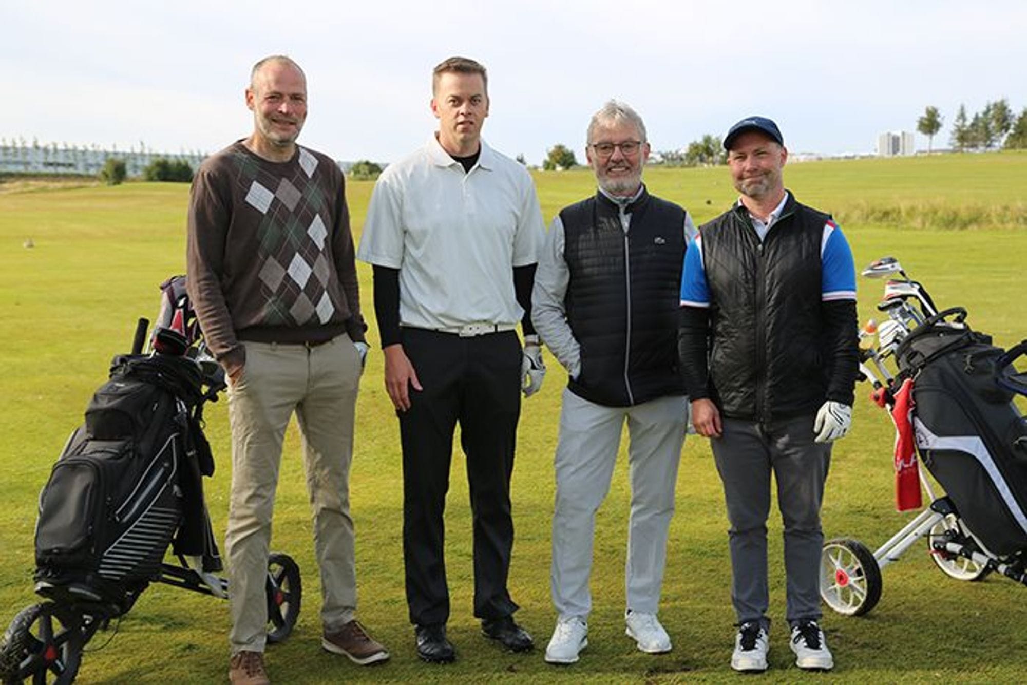 Four men standing together on the grass, under clear sky, with their golf equipment