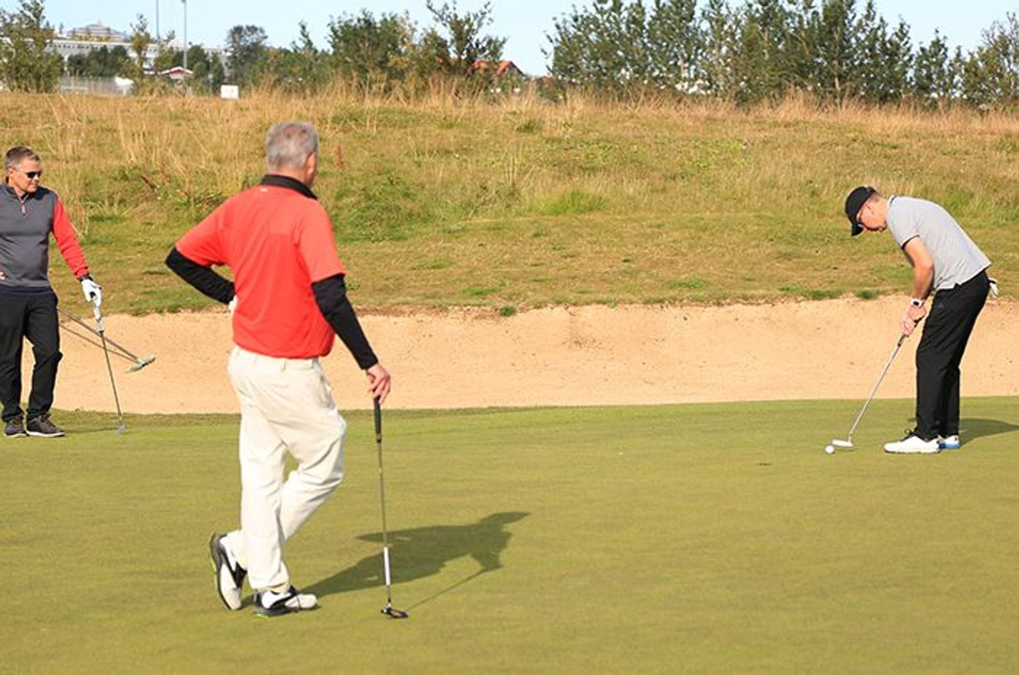 Three golfers are on a course with one preparing to putt and other two observing 