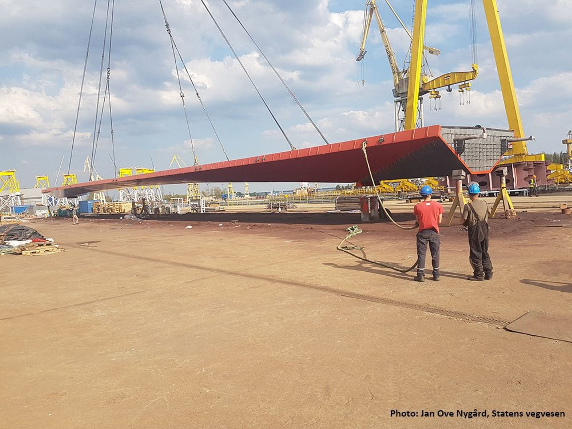 A large bridge segment being lifted by a yellow crane while workers observes it