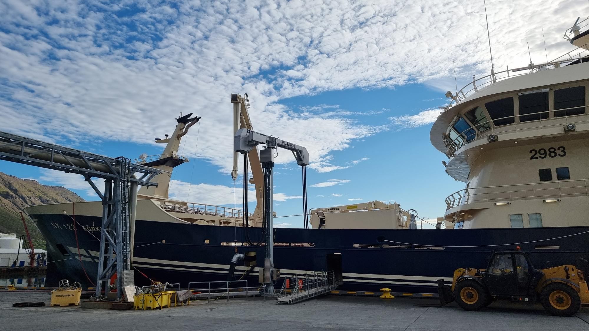 The photo captures a large fishing vessel, industrial equipment and a forklift under cloudy sky