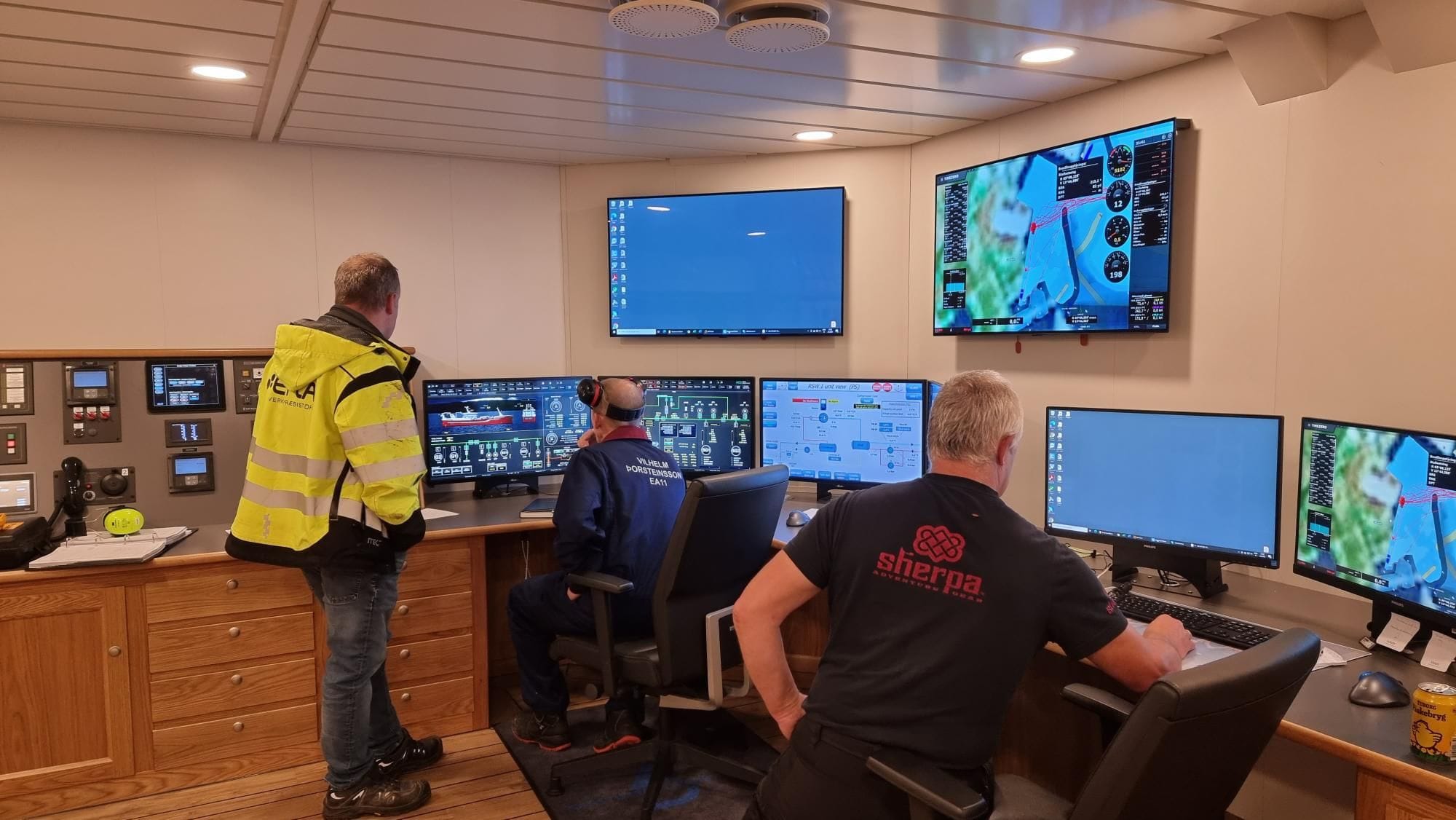 Three men in a control room with multiple computer screen displaying graphical interface and maps