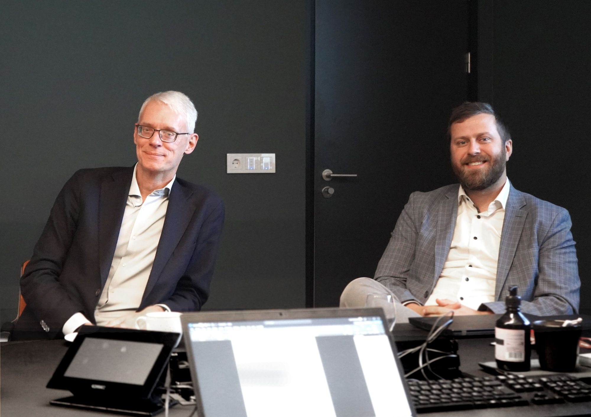 Two men in suits seated at a desk with laptops