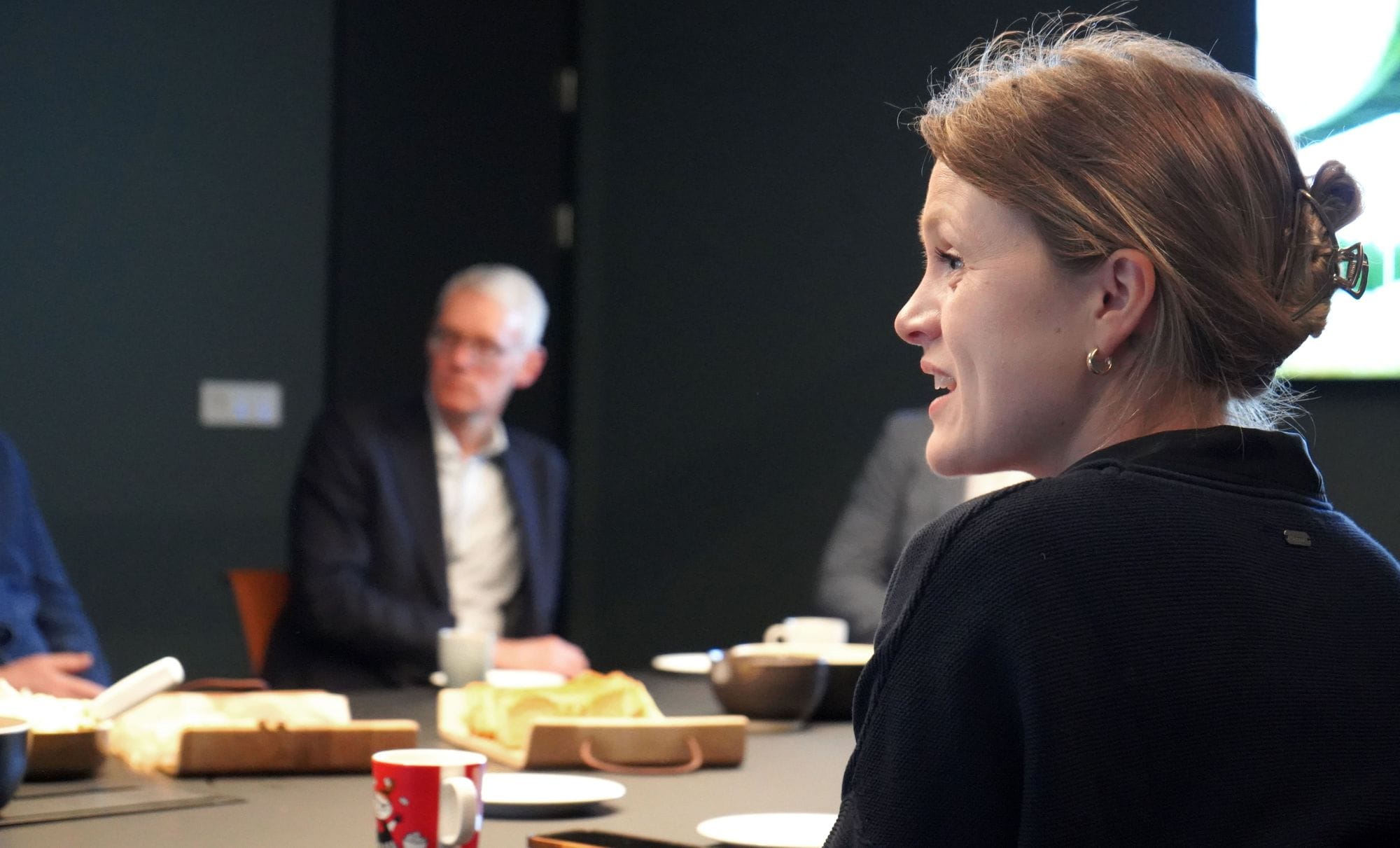 The photo captures a woman in focus, speaking or listening, and a blurred figure of a man in the background