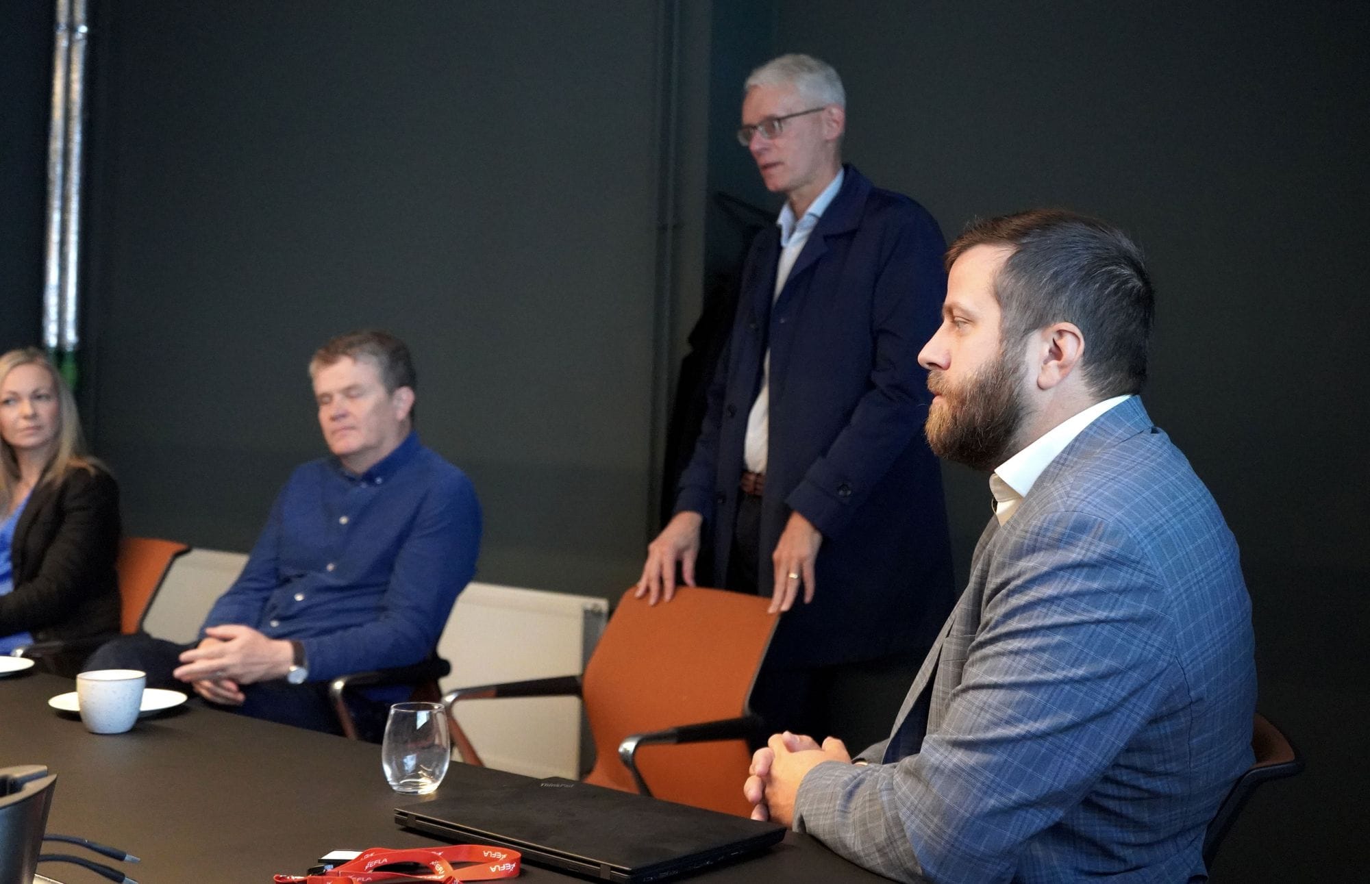 The photo captures four individuals in a meeting room seated and standing behind orange chair