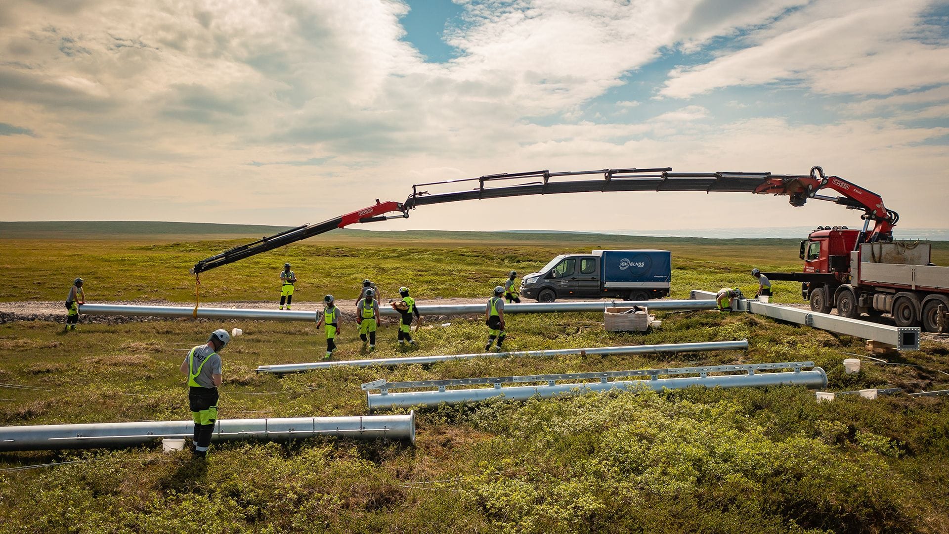 A construction site with workers in high visibility jacket, a crane, pipes on the ground and vehicles