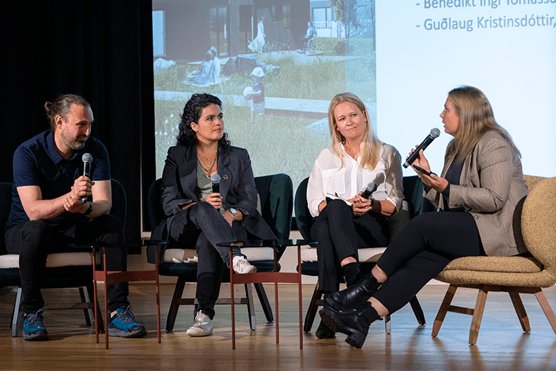 Four individuals seated with a big screen behind them, holding microphones and engaged in discussion 