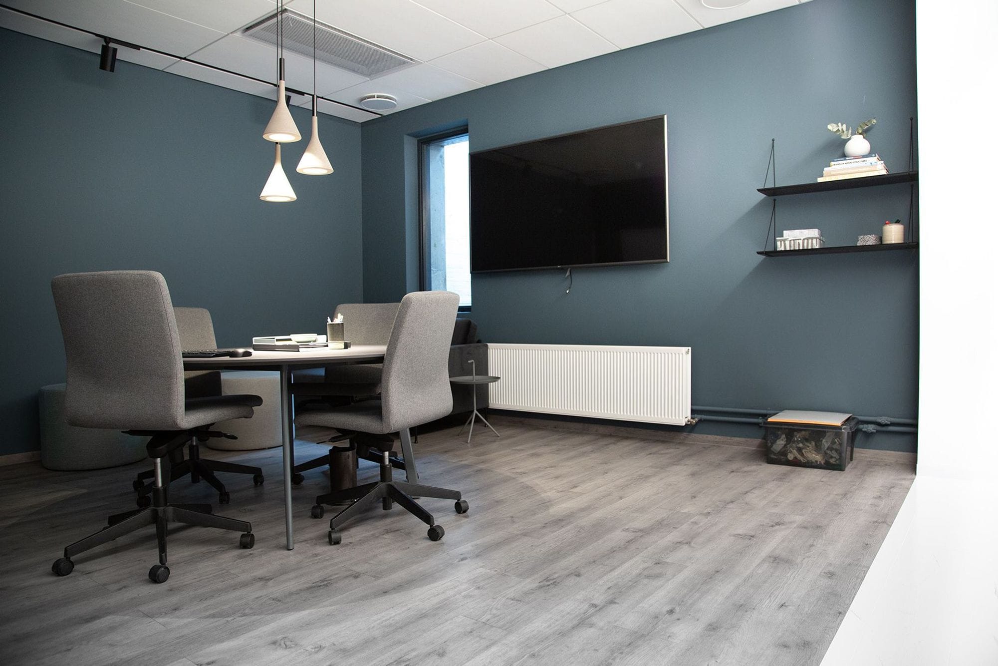 A room with a large round table , four grey office chairs, televisions on wall, pendant lights.