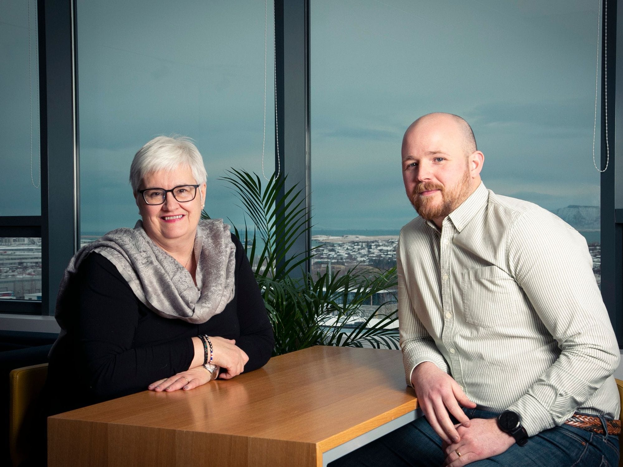 A man and a woman seated at a table inside an office with a view of a cityscape through the window behind them
