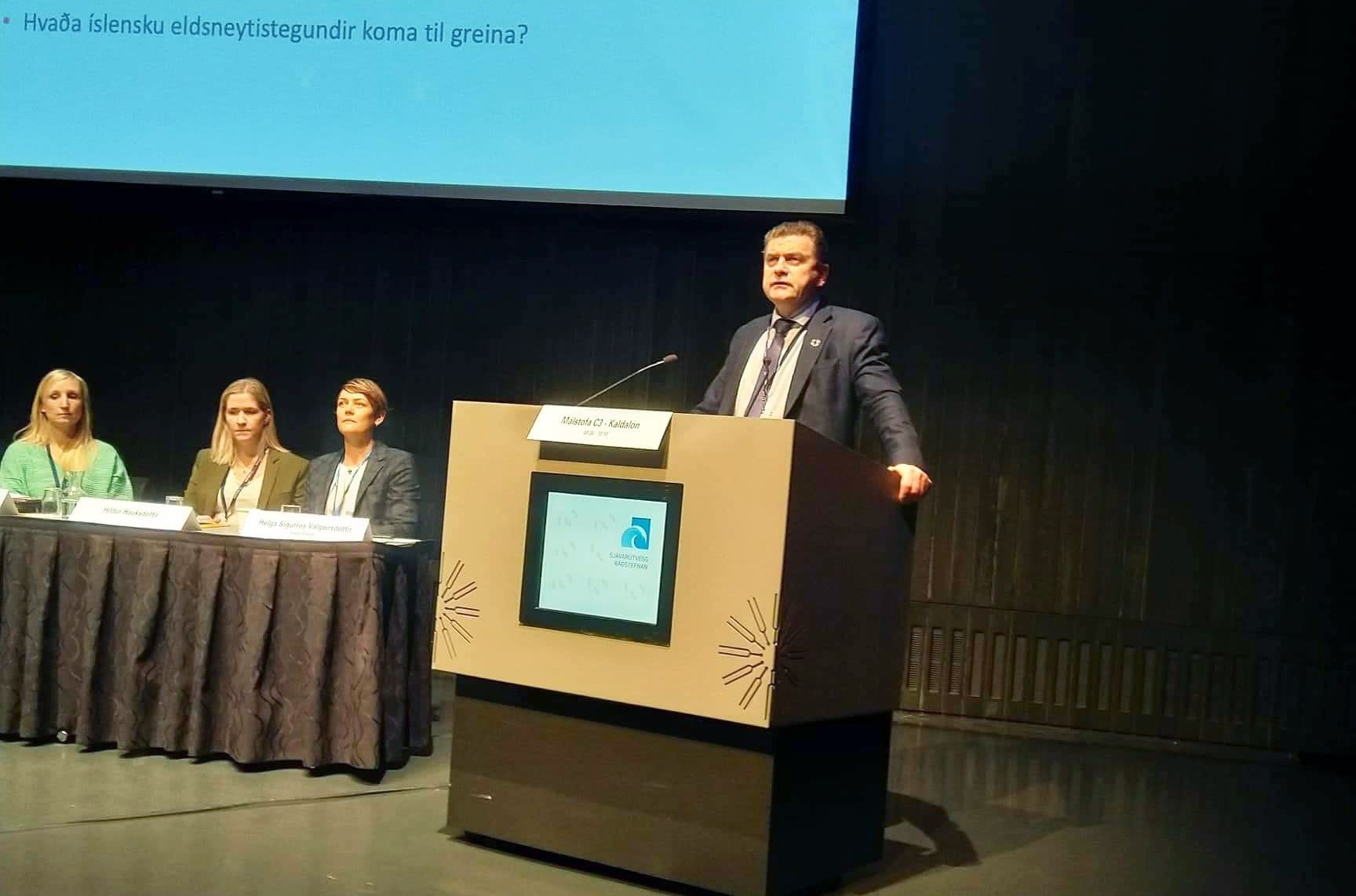 A man speaking at a podium with a panel of three women seated at a table behind him