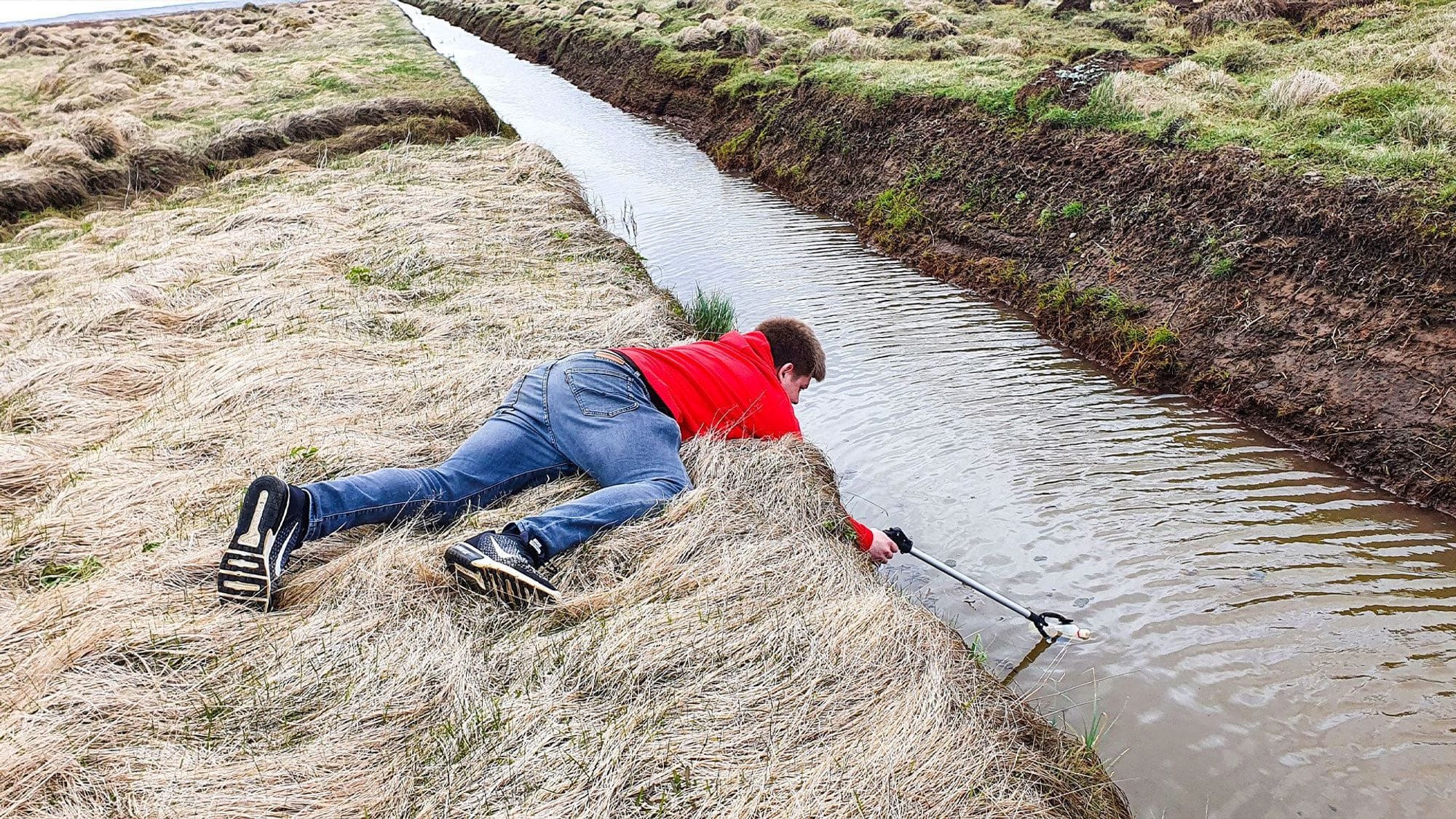 A person is lying on the ground reaching down into a ditch to catch a plastic container