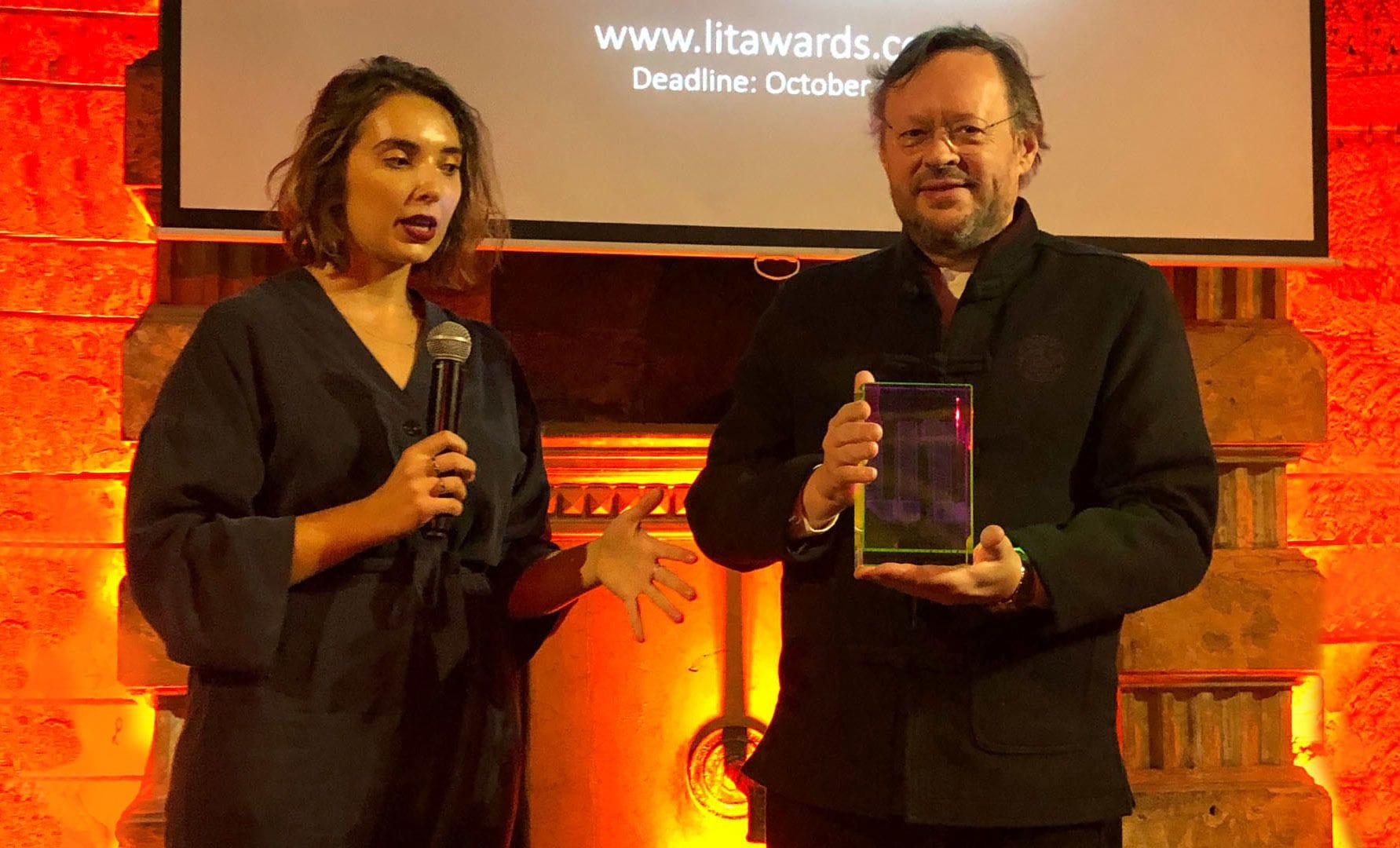A woman speaking into a microphone and a man holding an award next to her