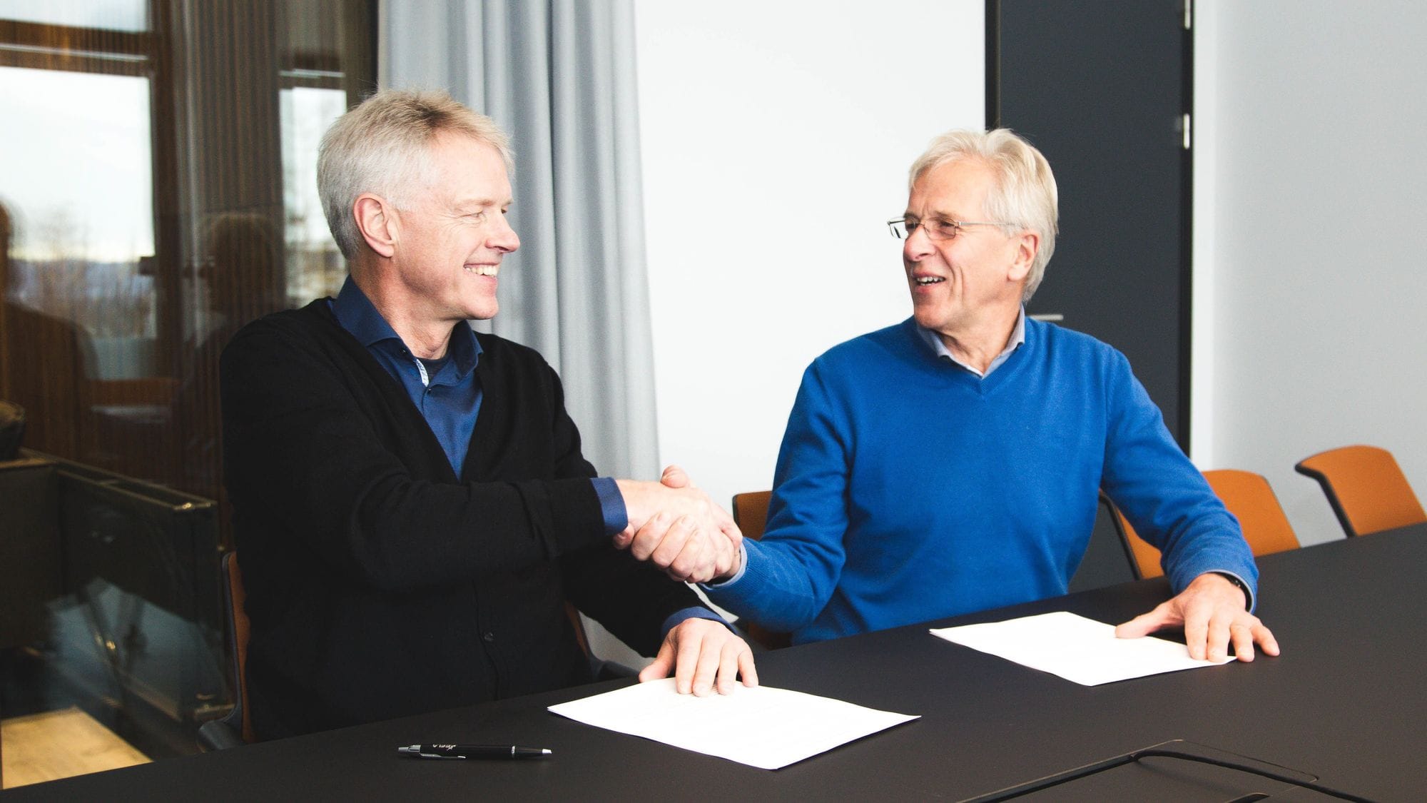 Two men seated, shaking hands with white paper on the table 