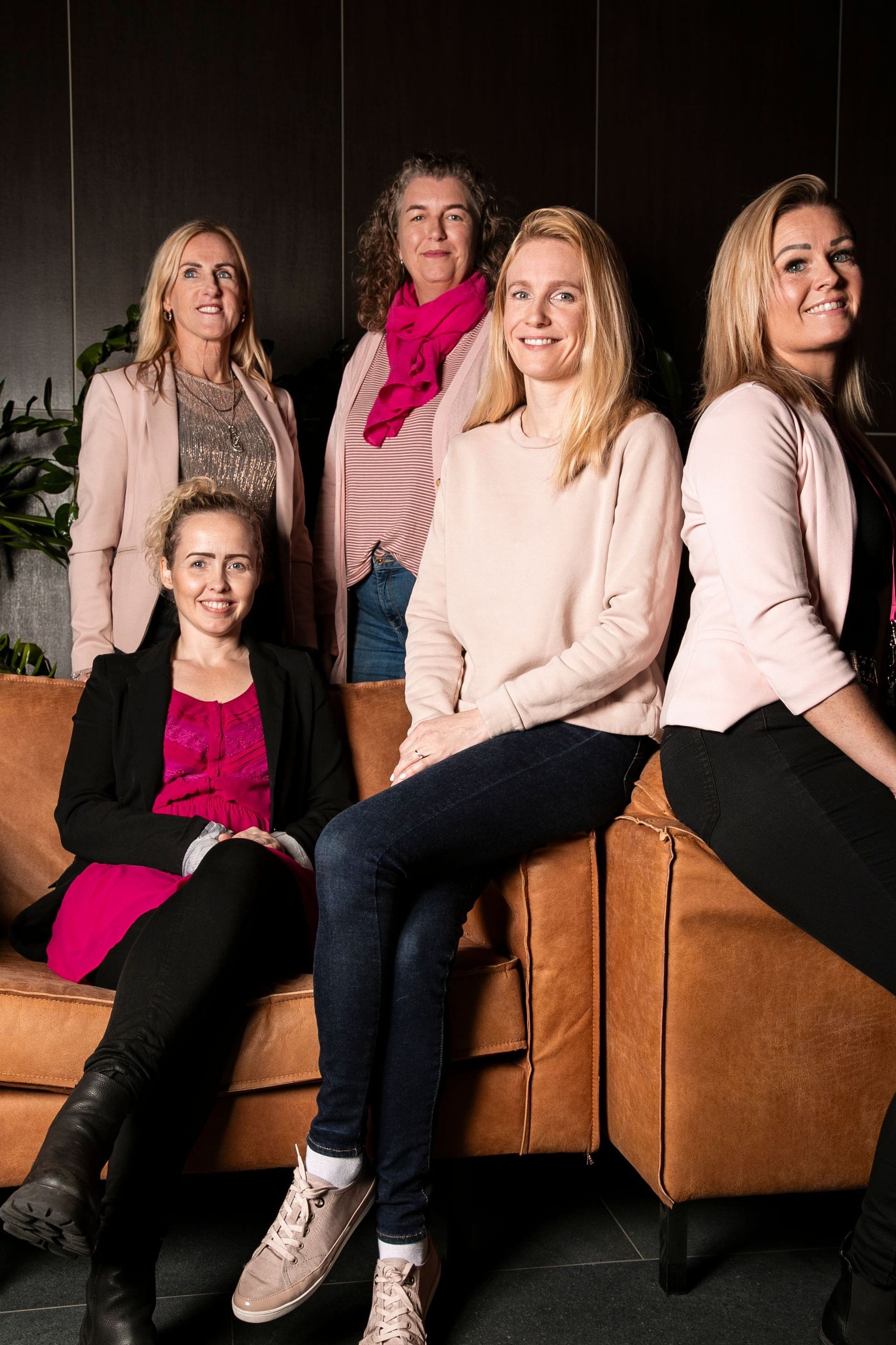 A group of five women, two standing and three seated on a leather couch, all set against a dark interior background with green foliage