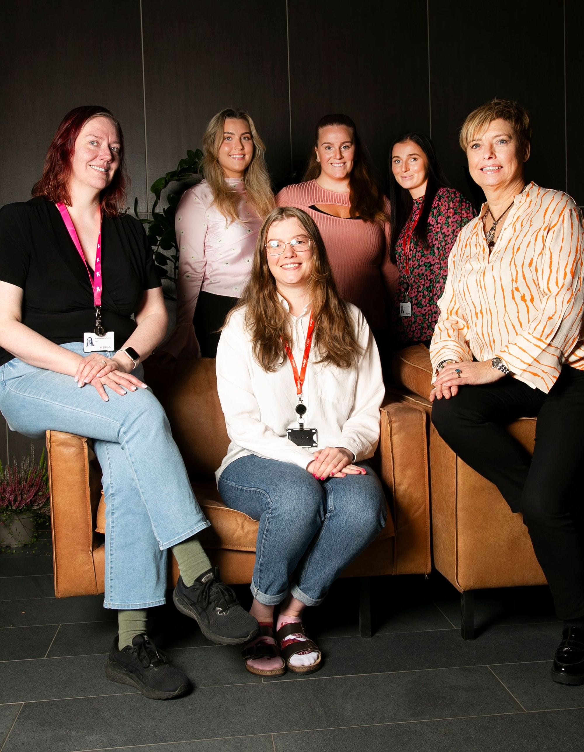 A group of six women, three standing and three seated on a leather couch, all set against a dark interior background with green foliage