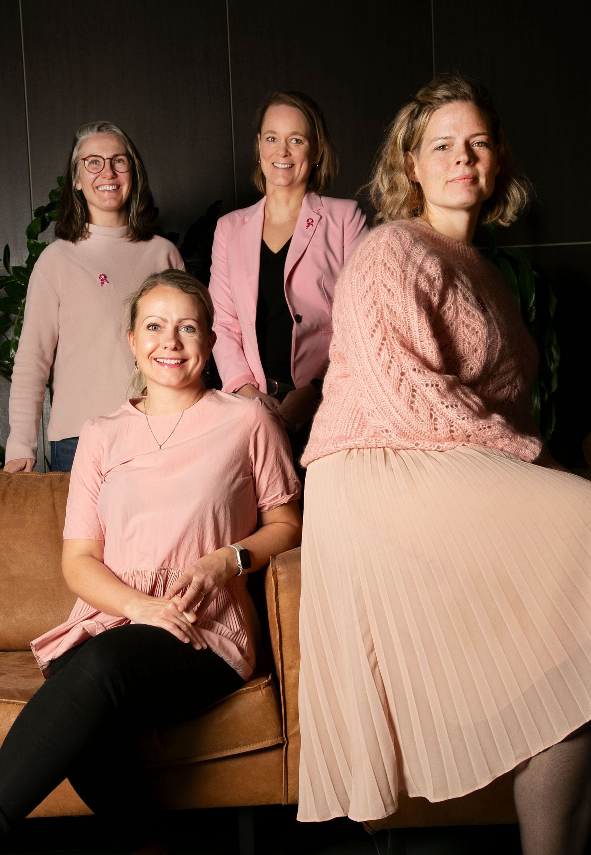 A group of four women, two standing and two seated on a leather couch, all set against a dark interior background with green foliage