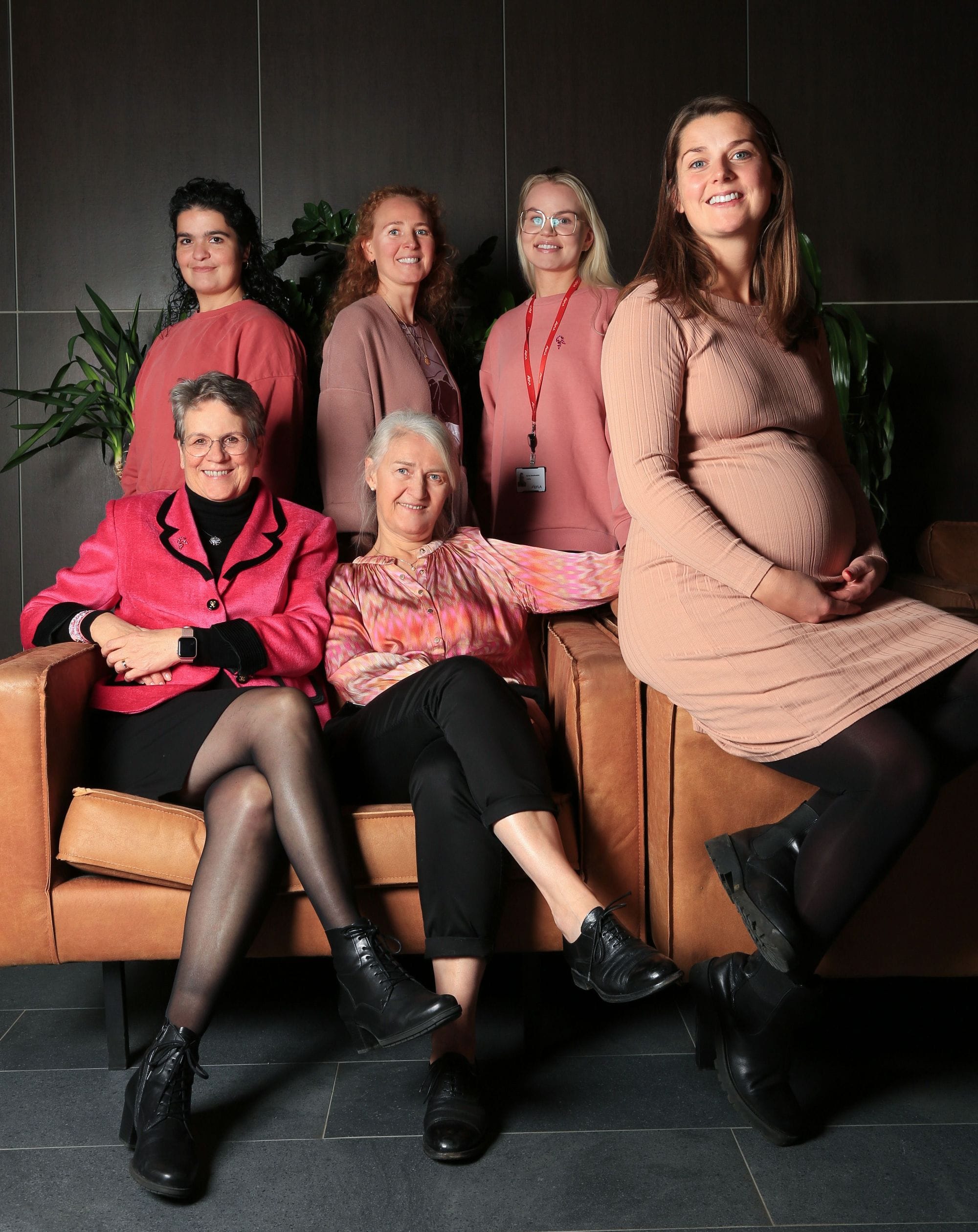 A group of six women, three standing and three seated on a leather couch, all set against a dark interior background with green foliage