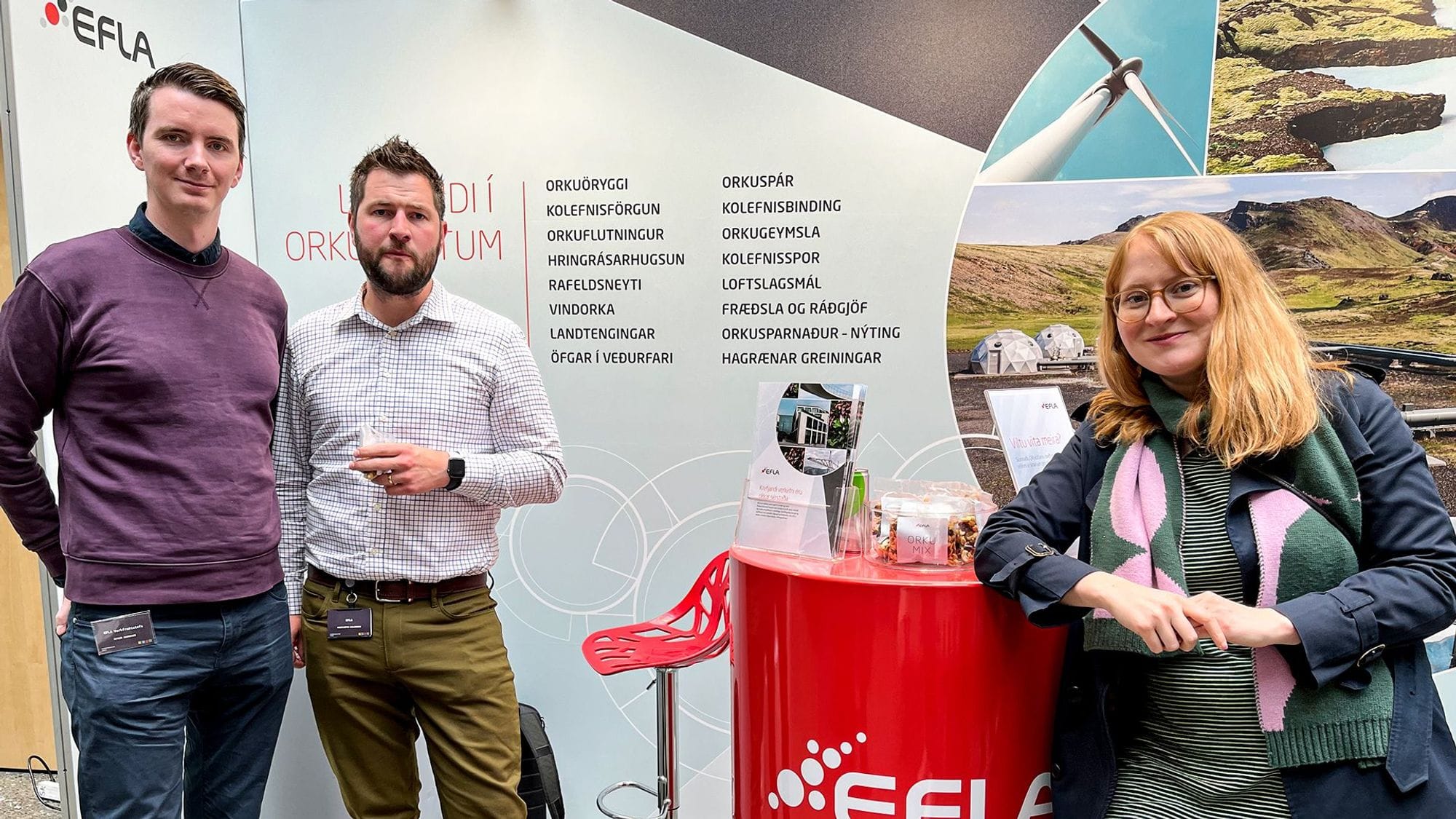 The photo features two men and a woman in front of a promotional stand, seemingly in an indoor corporate event or conference
