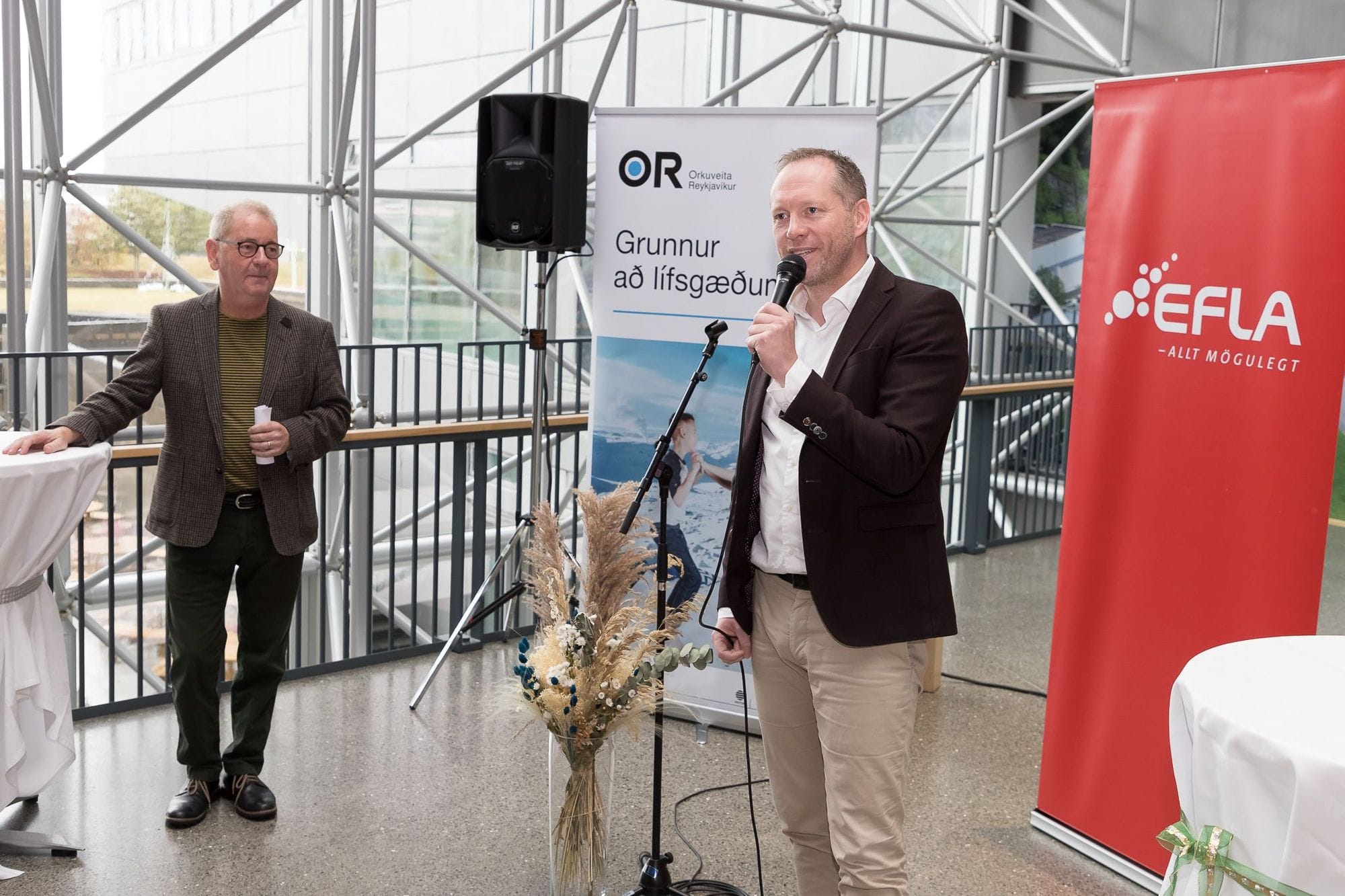 Two men in front of promotional banners, one speaking into a microphone and another one observing