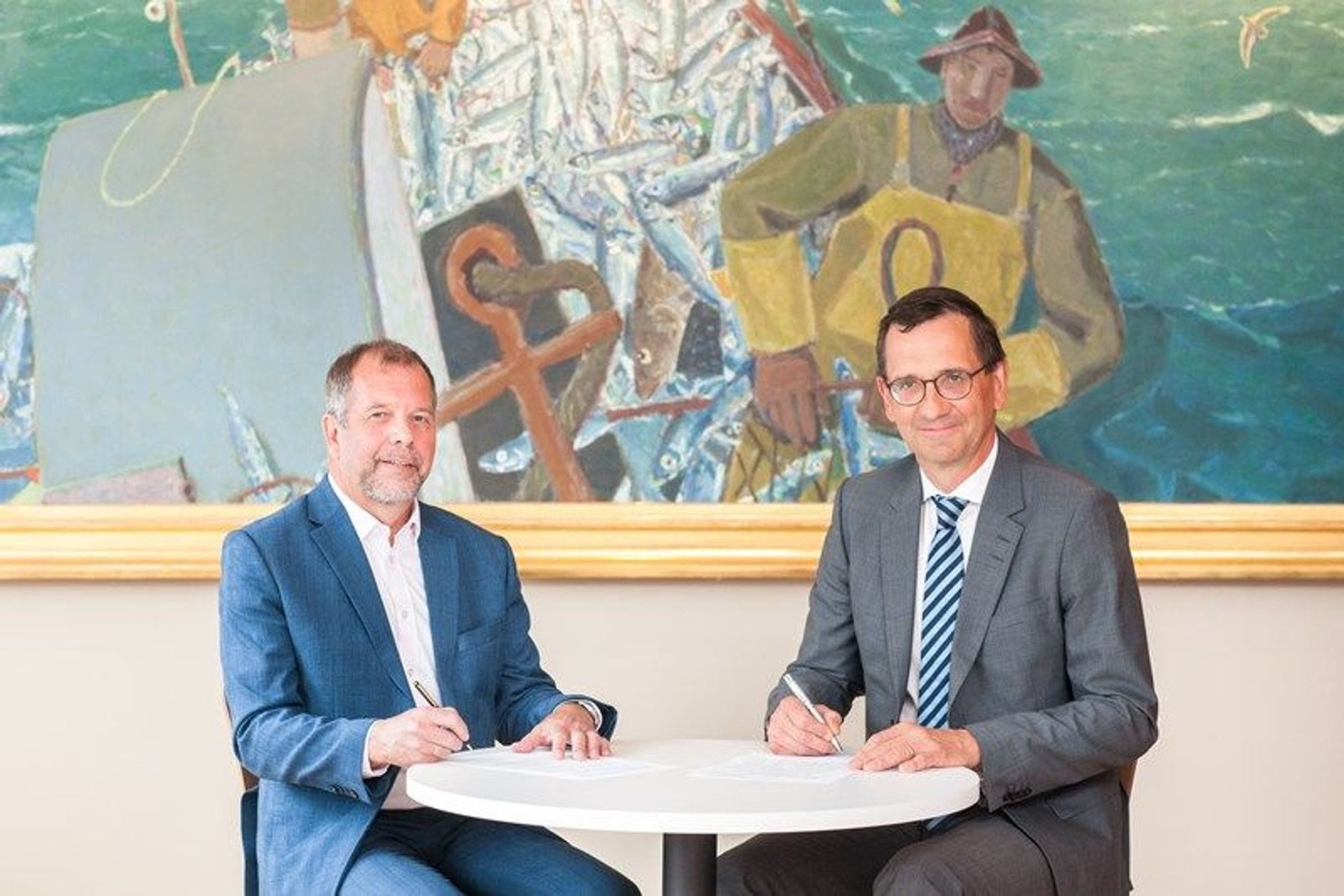 Two men seated around a round white table, writing on a white paper, with a fisherman painting in the background