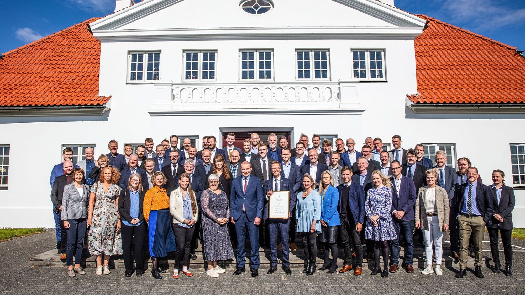 A large group of formally dressed people posing for a photo in front of a classic white building with a red tiled roof 